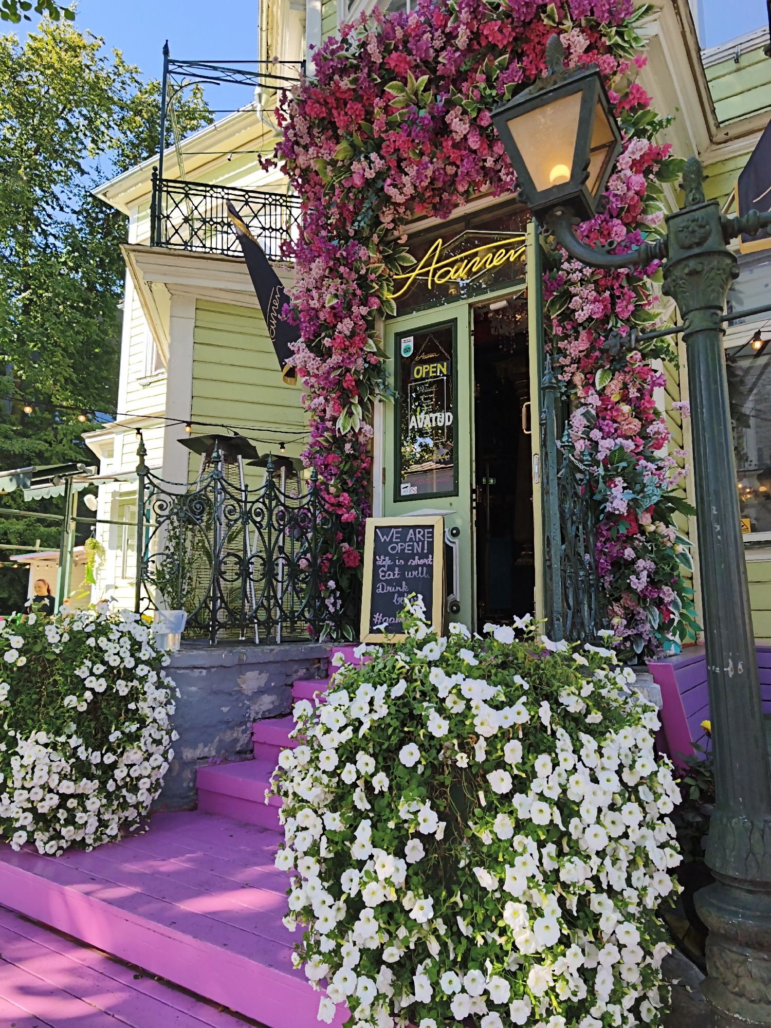 The image shows the exterior of the "Acuamen" restaurant in Tallinn, Estonia, on a sunny day. The entrance is adorned with an abundance of pink and purple flowers, creating a vibrant and welcoming facade. A neon sign above the entrance reads "Acuamen". To the left of the entrance, a green door is visible, and a sign on the door states "OPEN" in English and "AVATUD" in Estonian. A blackboard sign placed in front of the entrance reads "WE ARE OPEN! Life is short, Eat well, Drink well. #agfamm".
White petunias are abundant in large planters on either side of the pink-painted steps leading up to the entrance. A traditional black street lamp with a lit bulb stands to the right of the entrance. In the background, a balcony with an ornate railing is visible on the light green building, with a black flag hanging from it. Trees and greenery surround the building, and string lights are also present. The scene suggests a lively and attractive establishment in a city setting.