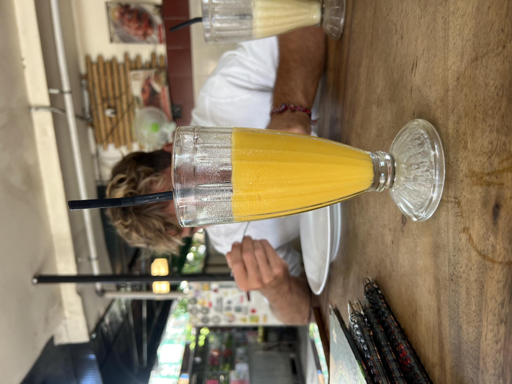 A close-up, eye-level shot shows a tall, fluted glass of bright orange juice with a black straw in the foreground, placed on a rustic wooden table. To the right, another glass containing a lighter-colored beverage is partially visible. A person with blonde, tousled hair, wearing a white t-shirt and a colorful woven bracelet on their right wrist, is blurred in the background, actively eating with a fork, with a white plate in front of them. The environment appears to be an open-air restaurant or cafe in Kuta, Indonesia, suggested by the tropical greenery visible through the open structure, awnings, and decorative wooden wall panels and artwork in the background. A stack of menus lies to the left of the orange juice glass. The lighting is bright, likely from natural daylight.