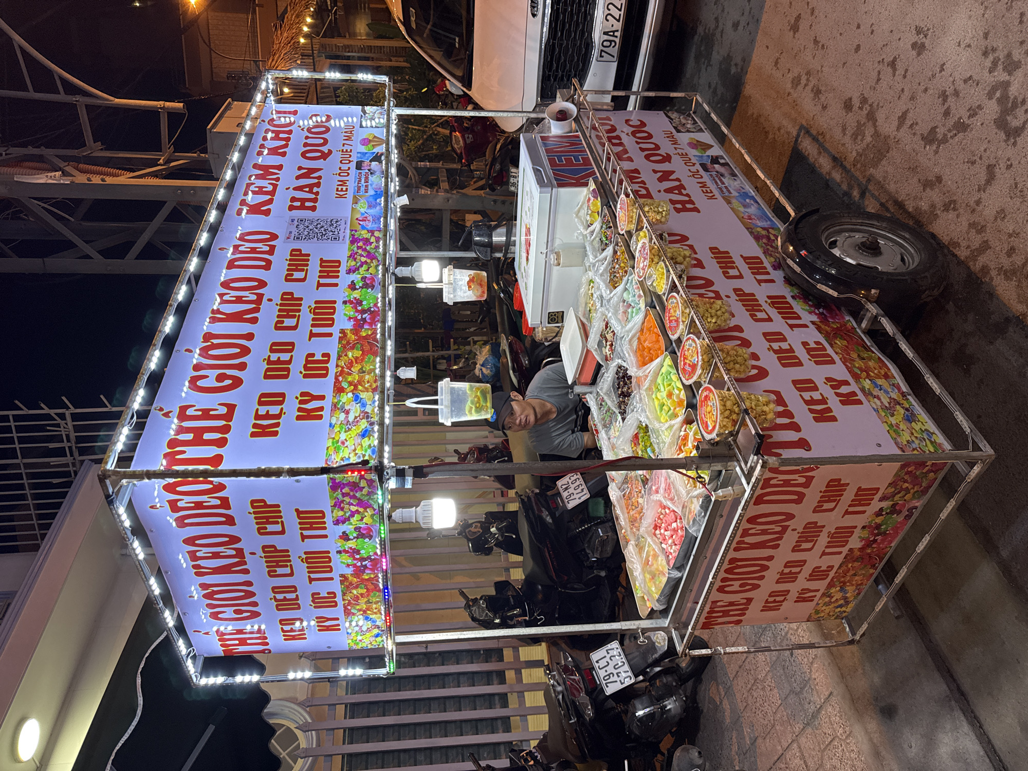 This image depicts a street food cart at night in Nha Trang, Vietnam. The cart is illuminated by strings of LED lights and features a large sign with Vietnamese text, advertising candy and ice cream. The cart is laden with various colorful candies displayed in clear plastic bags and containers. A man wearing a black cap and grey t-shirt is seated behind the cart, appearing to be the vendor. Several motorcycles are parked near the cart, and a white vehicle is partially visible on the right side of the image. The setting appears to be a street with a paved surface and a metal fence in the background. The overall scene suggests a busy evening street food stall.