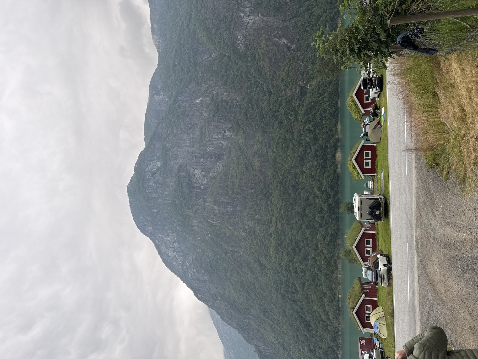 In Olden, Norway, a scenic camping area is depicted with a dramatic mountain backdrop. The foreground features a gravel road, with a paved road and grassy area separating it from a line of small, traditional red cabins topped with green, grassy roofs. Several tents are pitched on the grass in front of the cabins, along with parked cars and a camper van. The cabins appear to be part of a campground situated beside a serene, turquoise body of water. Beyond the water, a steep, densely forested mountain rises majestically, its rocky cliffs partially visible through the trees under an overcast sky. The overall scene conveys a peaceful, natural setting ideal for outdoor recreation.