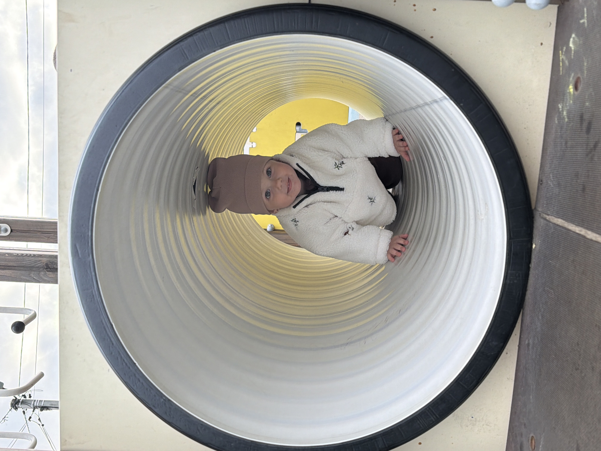 A child is inside a playground tunnel. The child is wearing a brown hat and a white fleece jacket. The tunnel is white with a black rim. The background shows a playground structure and sky. The location is Reshetnikovo, Russia.