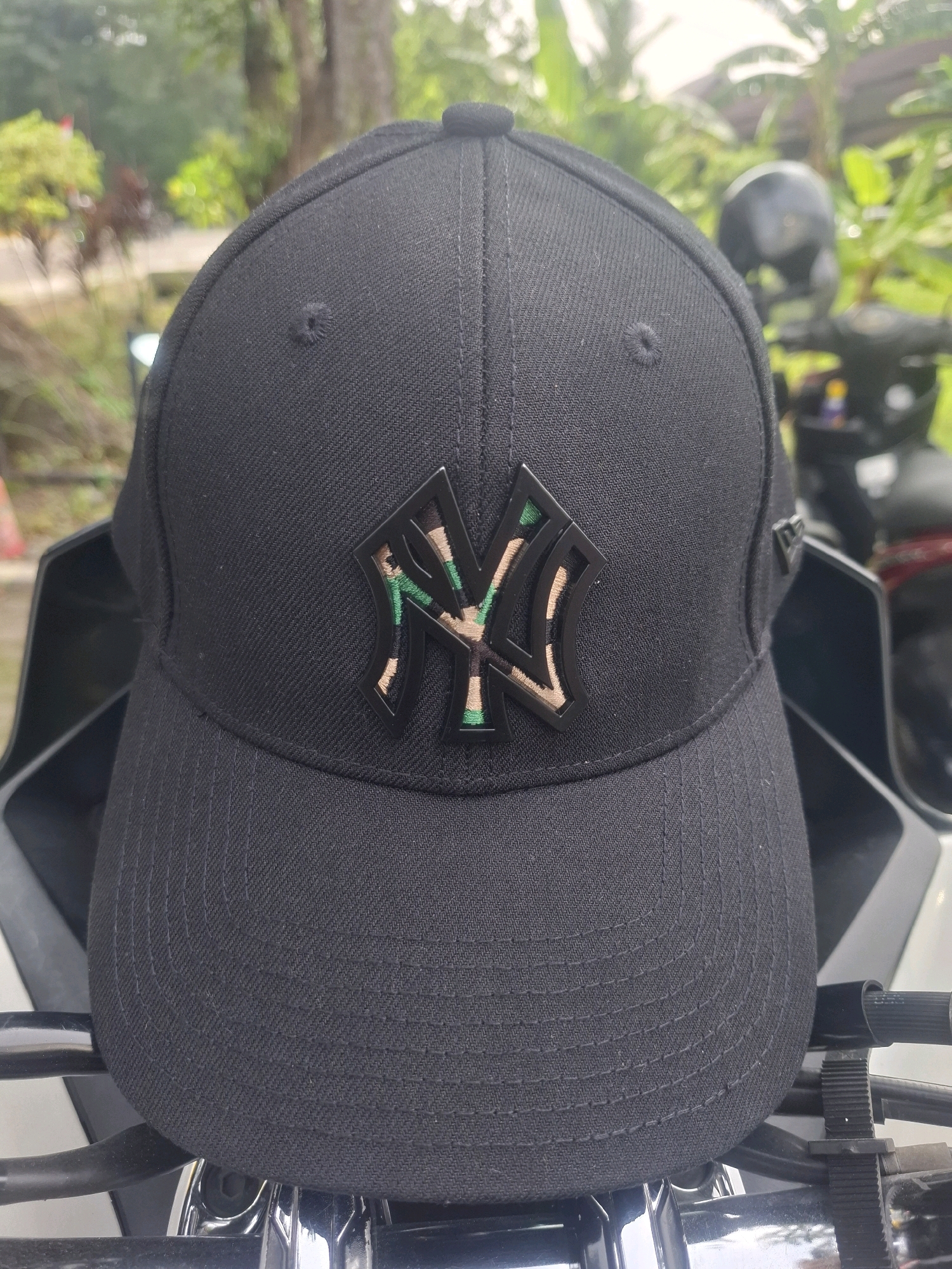 A close-up, eye-level, outdoor shot showcases a black New York Yankees baseball cap. The cap features a prominent black outline of the "NY" logo, with the interior filled with a camouflage pattern of tan, green, and light brown. The cap is positioned in the foreground, resting on what appears to be a motorcycle or similar vehicle, indicated by black handlebars and chrome accents visible below the cap. In the background, lush green foliage and trees are visible, suggesting a natural or park-like setting. The lighting suggests a bright, overcast day. The overall scene is static and focuses solely on the cap.