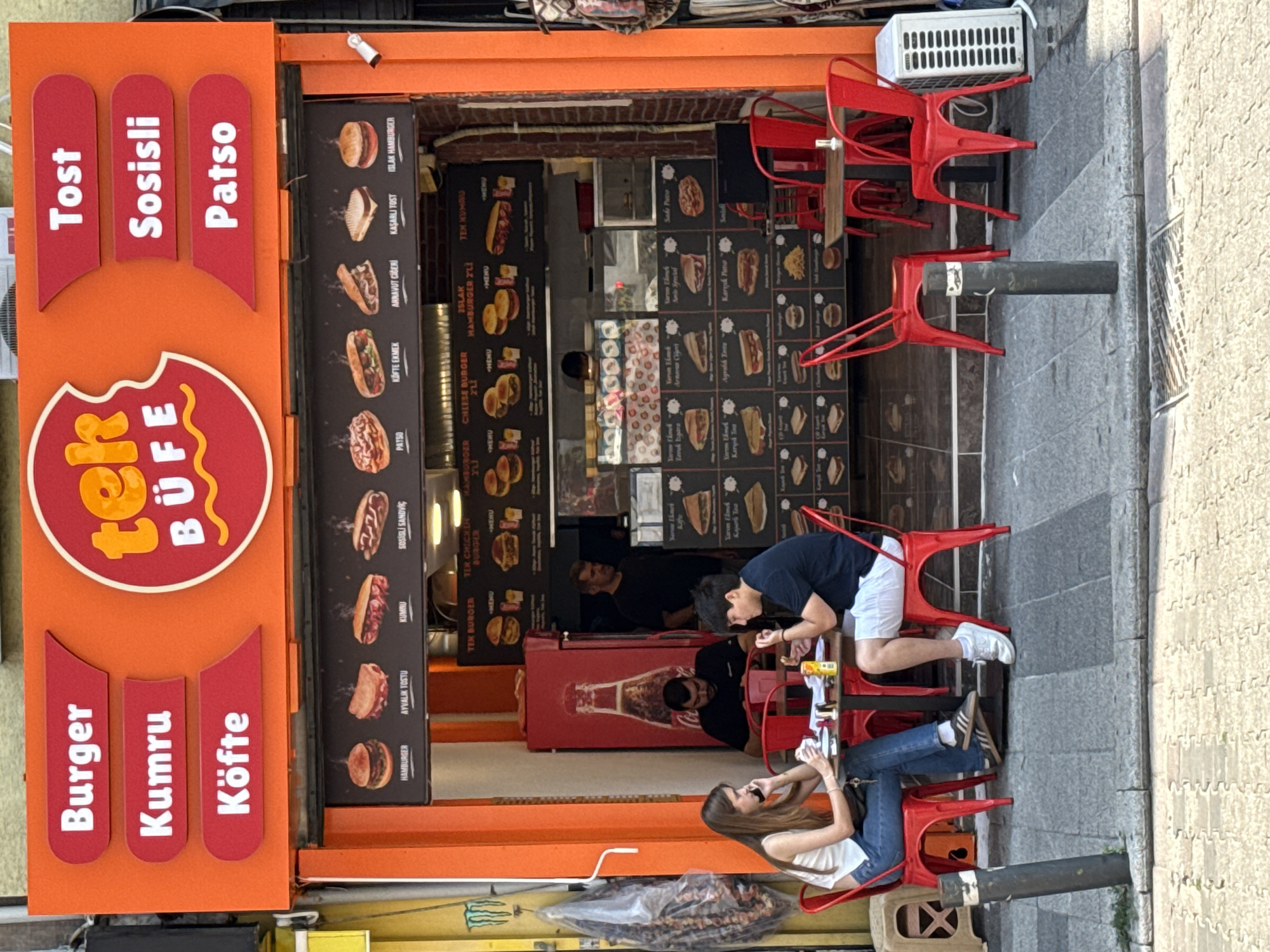 The image depicts a street-level view of "Tek Büfe," a fast-food establishment located in Kadıköy, Turkey. The facade is predominantly orange, featuring large red signage with white text listing menu items such as "Burger," "Kumru," "Köfte," "Tost," "Sosisli," and "Patso." A circular logo with the text "tek BUFE" is centrally positioned.
Below the main signage, a menu board displays images of various food items, including hamburgers, sandwiches, and Turkish specialties like "Kumru." The establishment has an open counter area where staff can be seen.
In the foreground, two young adults are seated at a small red table outside the shop. A woman with long brown hair, wearing a white sleeveless top and blue jeans, is talking on her phone and holding a piece of food. A man with dark hair, wearing a dark t-shirt and white shorts, is also seated at the table, holding a piece of food and looking downwards. Empty red chairs are visible around them, suggesting additional seating.
The ground is paved with grey interlocking stones and concrete. A black bollard stands in the foreground, and further back, a white air conditioning unit is placed against the building. The overall scene is typical of a casual street-side eatery in a bustling urban area.