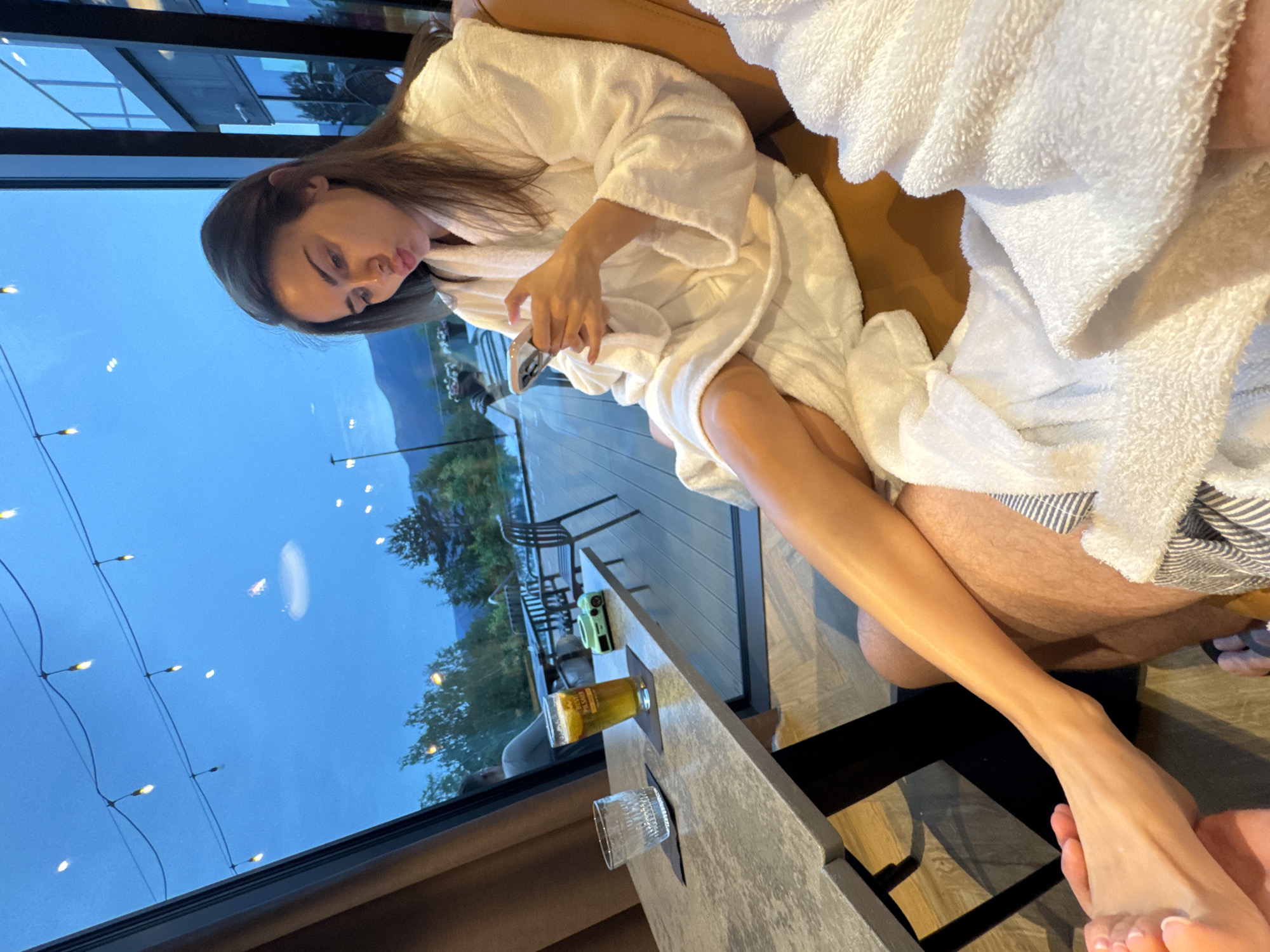 An indoor scene captured during dusk or early evening shows two individuals relaxing by a large window overlooking an outdoor area and distant mountains. A woman with long dark hair, wearing a white bathrobe, is seated comfortably on a brown sofa, holding a smartphone in her hands and looking at it with a slightly pouting expression. Her right leg is extended.
Next to her, a man is partially visible, also wearing a white bathrobe and striped fabric (possibly shorts or a towel). His legs are hairy. On a small table in front of them are a glass containing a light amber drink, likely beer, with a slice of citrus, and an empty clear glass. The beer glass has "BEST PILSNER" written on it. A green instant camera also sits on the table.
The large window reveals a blue twilight sky with string lights illuminated outside. Beyond the outdoor deck, trees and the silhouette of mountains are visible. The atmosphere is calm and relaxed, suggesting a leisure or spa setting. The woman's right foot is seen in the foreground, being gently held, possibly by the man's foot.