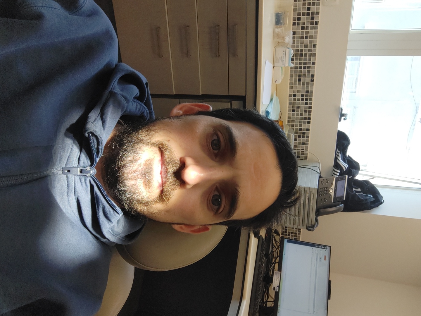 A selfie shot features a man with dark hair and a trimmed beard. He's wearing a zipped-up blue hoodie and seated in what appears to be a beige dental chair. The background includes office elements: a tiled backsplash, drawers, a computer monitor, a keyboard, a telephone, and a window. Outside the window, sunlight streams, although specific landmarks indicating Tel-Aviv, Israel, are not visible.