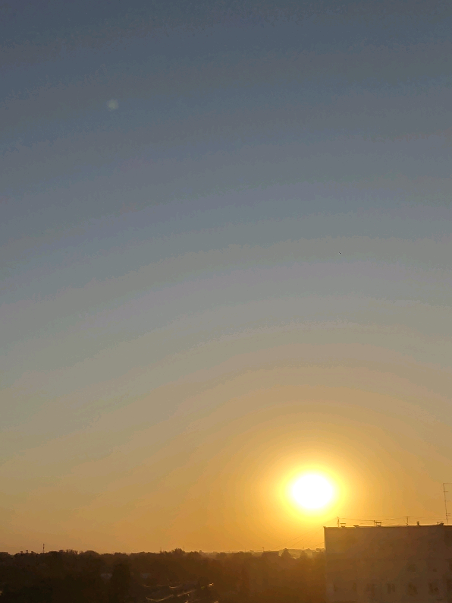 A wide shot captures a sunset over Rubtsovsk, Russia. The sun, a brilliant orb of light, is positioned in the lower right quadrant of the frame, casting a warm, golden glow across the sky. The sky transitions from a pale blue at the top to a vibrant orange and yellow as it approaches the horizon. In the distance, the silhouette of the city is visible, with buildings and trees forming a dark, linear shape against the colorful sky. The foreground is dominated by the dusky silhouettes of trees and some residential buildings. One building, a multi-story structure, is prominently featured in the lower right corner, its upper edges catching the last rays of sunlight. The overall scene depicts a peaceful evening in the city, with the natural beauty of the sunset dominating the view. There are no people or animals visible in the image.