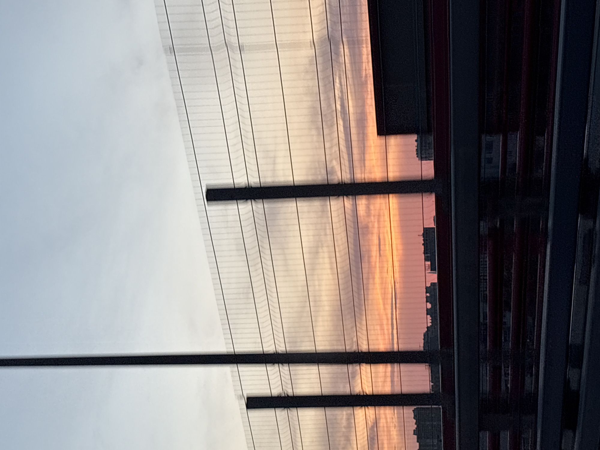 The image depicts a city skyline at dusk, viewed through a wire fence. The sky is a gradient of pale blue at the top, transitioning to soft orange and pink hues closer to the horizon, indicative of a sunset or sunrise. Silhouetted buildings of varying heights are visible in the distance, suggesting an urban environment. In the foreground, a tall, dark pole stands vertically, partially obscuring the view of the sky and fence. The fence itself is a mesh of horizontal wires and vertical supports. Below the fence, a dark structure with red horizontal elements is present, possibly a bridge or overpass railing. The overall impression is one of an urban landscape at twilight, framed by man-made structures.