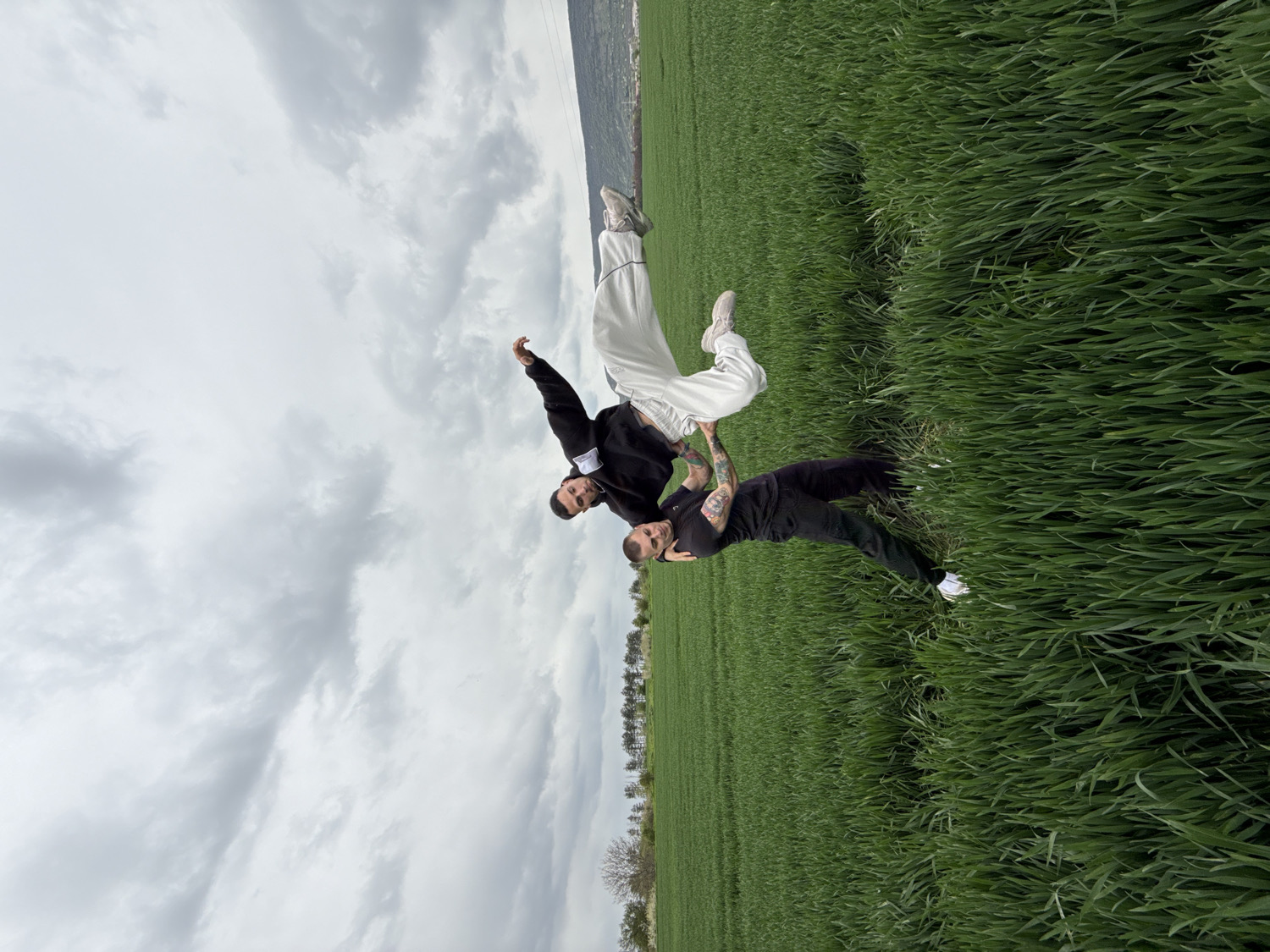 Two men are engaged in an acrobatic pose in a vast green field, under an overcast sky. One man, positioned at the bottom, wears a black long-sleeved top, black pants, and white sneakers, with visible tattoos covering his forearms. He is supporting the second man, who is inverted, balancing on the first man with his head pointing downwards and legs extended upwards. The inverted man wears a black short-sleeved shirt, loose white pants, and white sneakers. Both individuals appear focused on maintaining the delicate balance of the stunt.
The setting is a sprawling agricultural field, densely covered with vibrant green crops, likely young wheat or another grain, stretching towards a distant horizon. The sky is entirely covered with thick, grey clouds, indicating an overcast day with diffused light. A faint line of distant trees or hills is visible at the horizon line in the background. A small, white rectangular patch with dark, illegible text is visible on the left sleeve of the man in the black long-sleeved top. The scene takes place outdoors in Shumen, Bulgaria, capturing a moment of physical strength and coordination within a natural landscape.