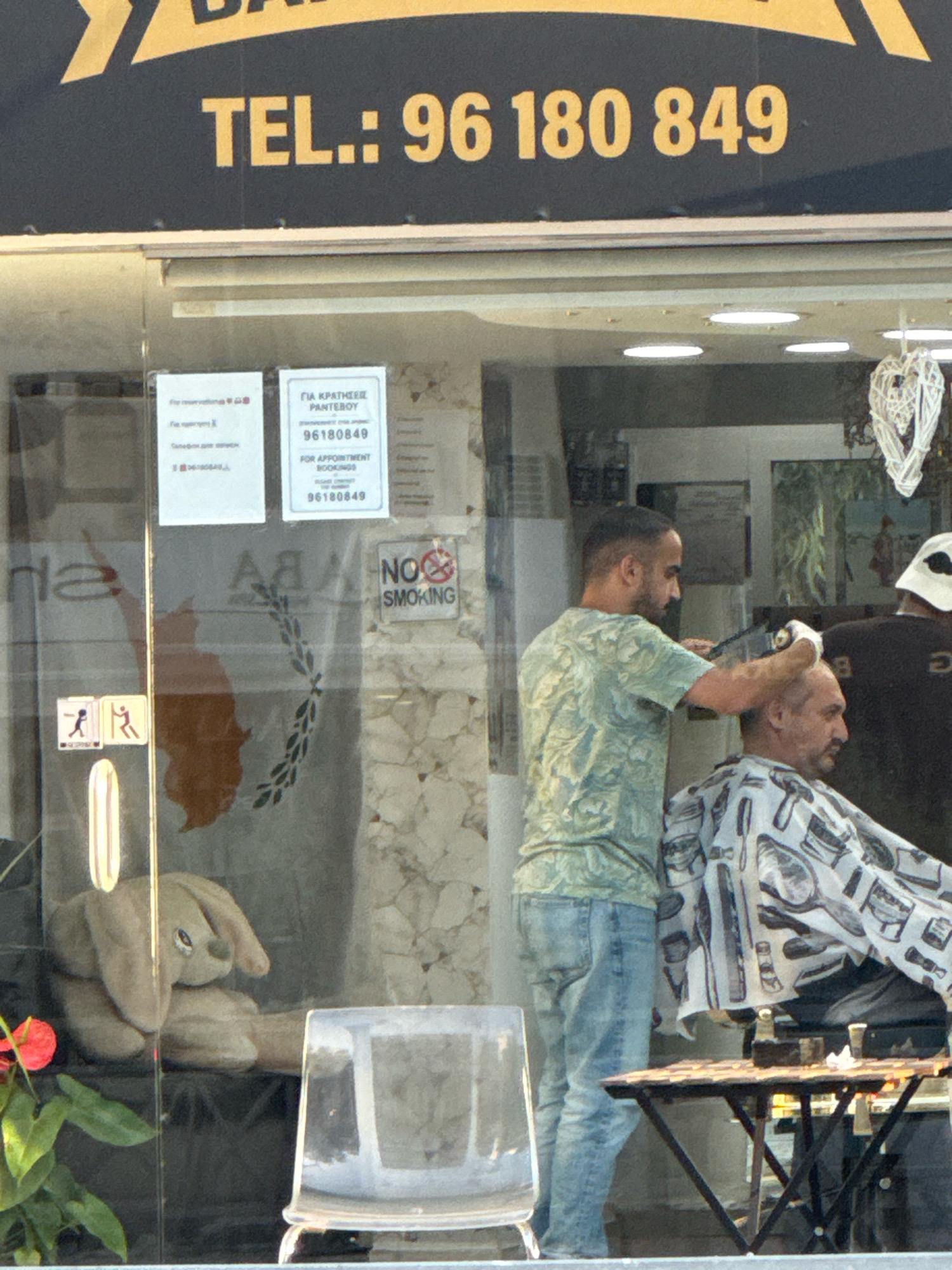 The image depicts a barber shop, likely located in the city of Germasogeia Municipality, Cyprus. A barber, wearing a green and white floral patterned shirt and blue jeans, is in the process of cutting the hair of a seated customer. The barber is holding clippers in his right hand and appears to be focused on the task. The customer, who is wearing a cape with a barber-themed pattern, is looking away from the camera. In the background, a second barber or customer is partially visible, wearing a white hat. Signs are displayed on the glass storefront, including one with the text "TEL.: 96 180 849" and another in Greek and English stating "ΓΙΑ ΚΡΑΤΗΣΕΙΣ PANTEBOY 96180849 FOR APPOINTMENT BOOKINGS 96180849". A "NO SMOKING" sign is also visible. Inside the shop, a stuffed rabbit toy is placed near the window, and a decorative heart ornament hangs from the ceiling. A small table with items on it is positioned in front of the seated customer. The overall environment suggests a functional and active barber shop.