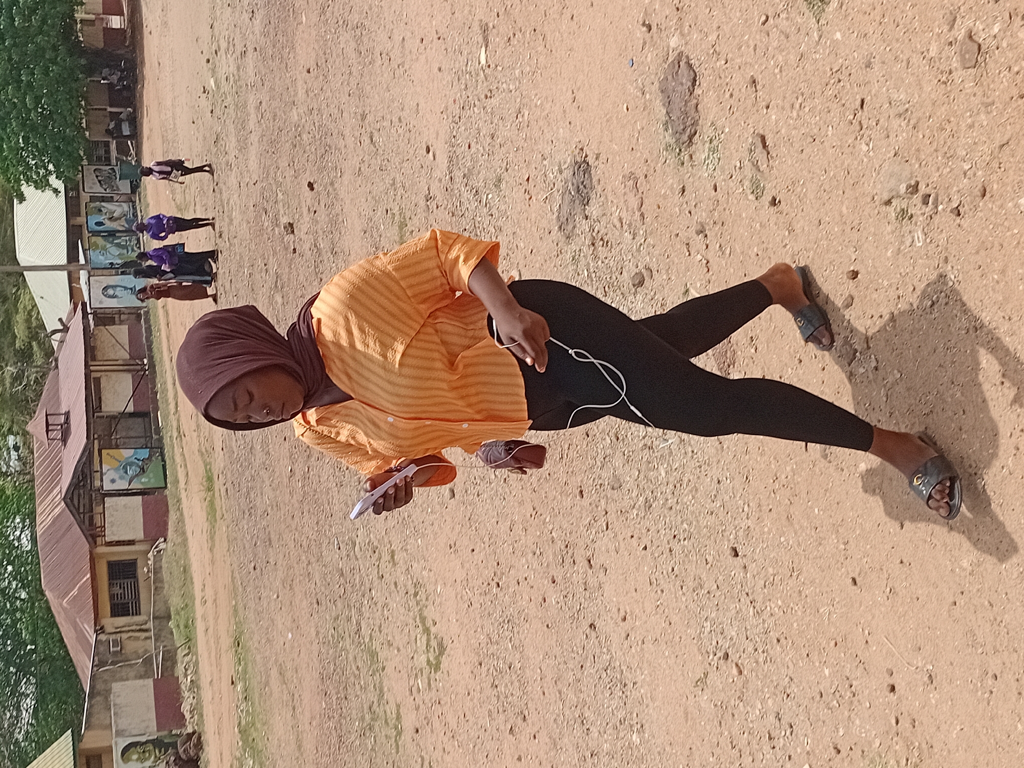 A woman stands outdoors in what appears to be a schoolyard in Lokoja, Nigeria. She wears a brown hijab, an orange striped shirt, and black leggings. She holds a white cell phone in one hand and white earphones are dangling in the other. She wears sandals. Behind her, a building with murals is visible, and in the distance, there are figures in purple shirts. The ground is arid and sandy.
