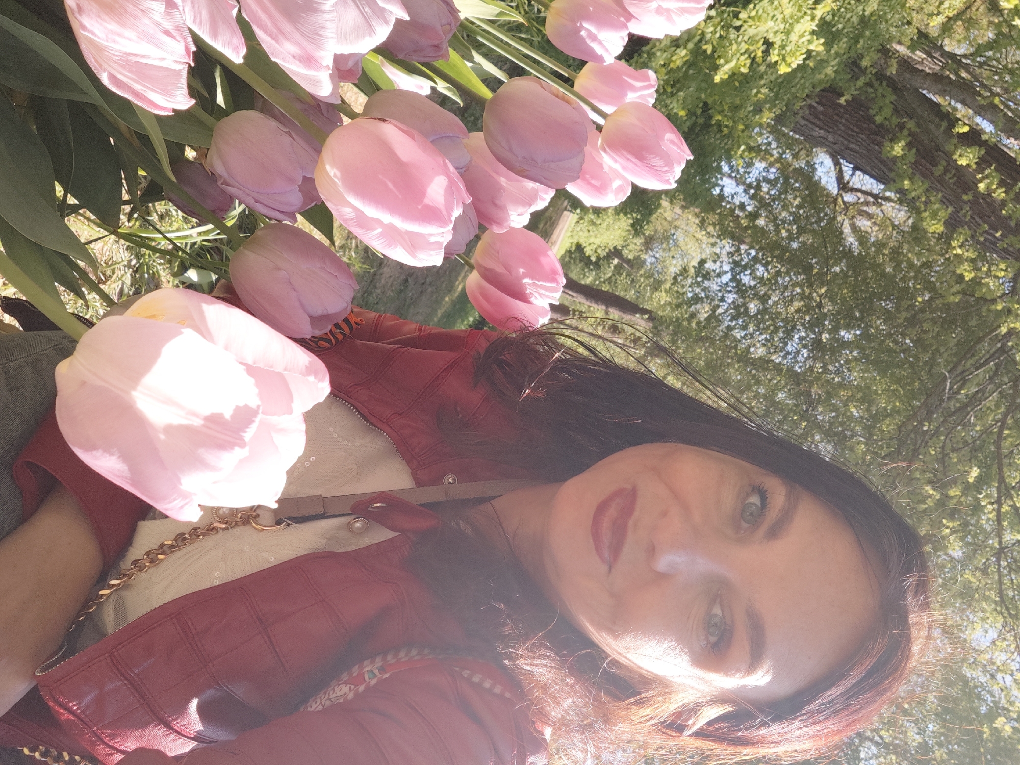 A woman is outdoors, likely in a park or garden, judging from the trees and flowers visible in the background. She is surrounded by numerous light pink tulips. The woman is wearing a red leather jacket over a white top with what appears to be a gold chain detail. The location is most likely one of the parks or gardens in the city of Saint Petersburg, Russia.