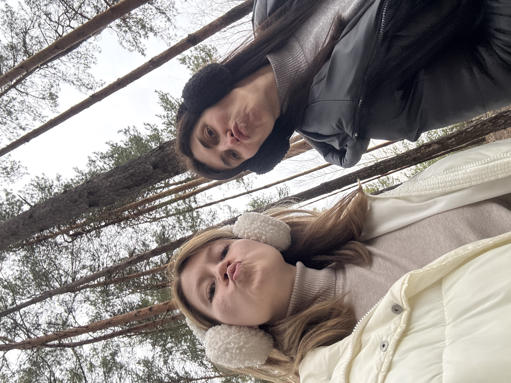 Two women are posing for a selfie outdoors, seemingly in a forest or wooded area. The photo is taken from a low angle, looking up at the subjects and into the trees above.
The woman on the bottom of the frame has blonde hair and is wearing white earmuffs and a white puffer jacket over a beige turtleneck sweater. She is puckering her lips as if to give a kiss.
The woman above her has dark hair and is wearing black earmuffs and a dark-colored jacket over a gray sweater. She is also puckering her lips.
Bare tree trunks and branches are visible in the background, creating a dense network against a light sky. The scene suggests a cold or winter environment, with the women dressed in warm clothing.
There is no clear evidence indicating the location is in the city of Chaikovsky, Russia. The presence of a forest and two women dressed in warm clothing does not specifically imply any unique characteristic of Chaikovsky.