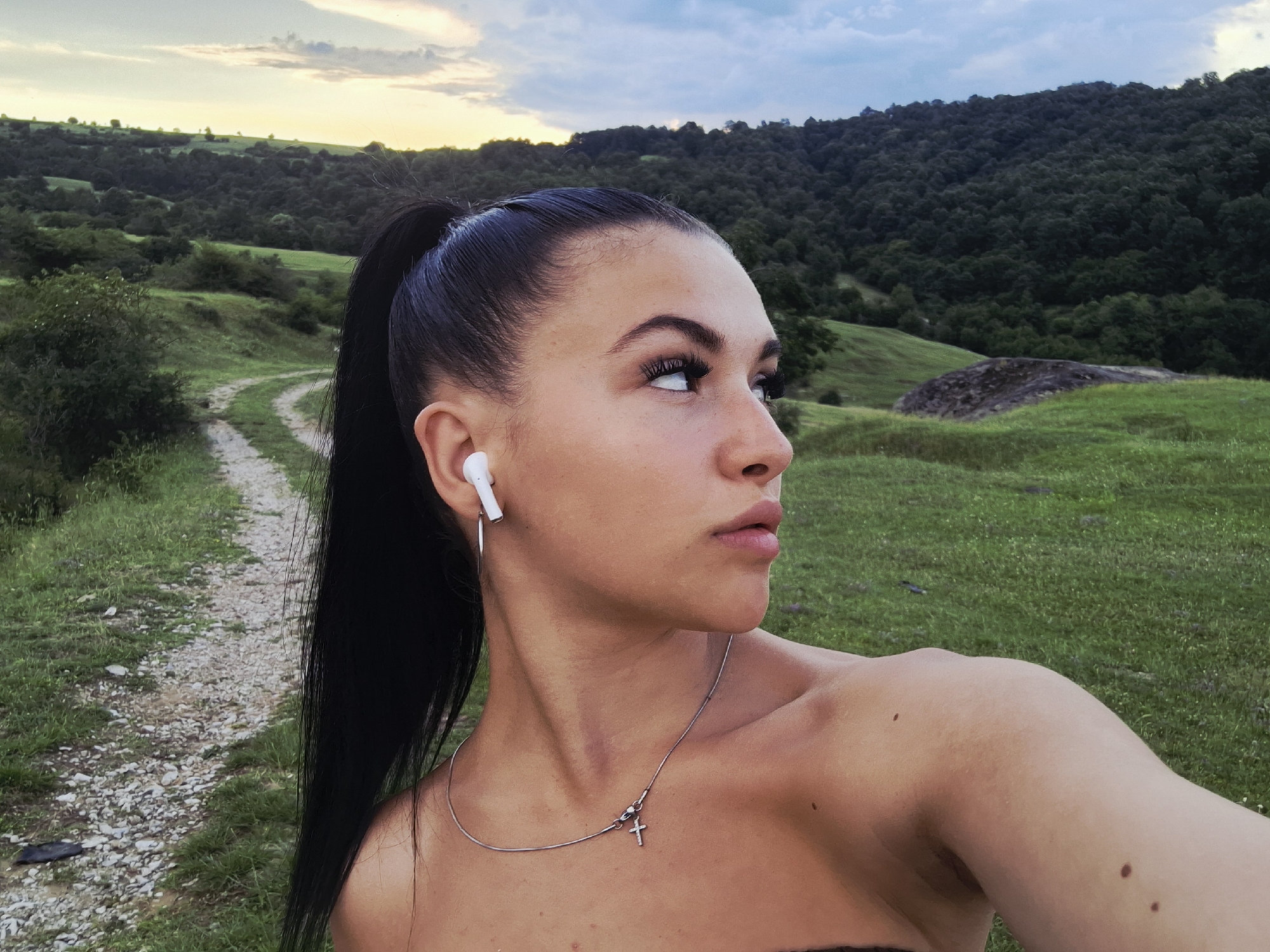 A young woman is captured in a close-up outdoor portrait, likely a selfie, looking intently towards her right with a neutral or contemplative expression. She has dark hair styled in a high ponytail and wears a white wireless earbud in her visible left ear. A silver necklace with a small cross pendant adorns her neck. Her bare shoulder and arm show a few moles.
The setting is a picturesque natural landscape, characterized by rolling green hills covered in grass, bushes, and dense forestation in the mid-ground and background. A winding dirt path meanders through the grassy terrain to the left of the frame. To the right, a large, dark rock or mound rests on a green slope. The sky above is partly cloudy, displaying soft blue tones with hints of warm yellow and orange light on the horizon, indicative of late afternoon or early evening. The overall scene conveys a sense of tranquility and natural beauty. The location is Krapets, Bulgaria. There is no visible text in the image.