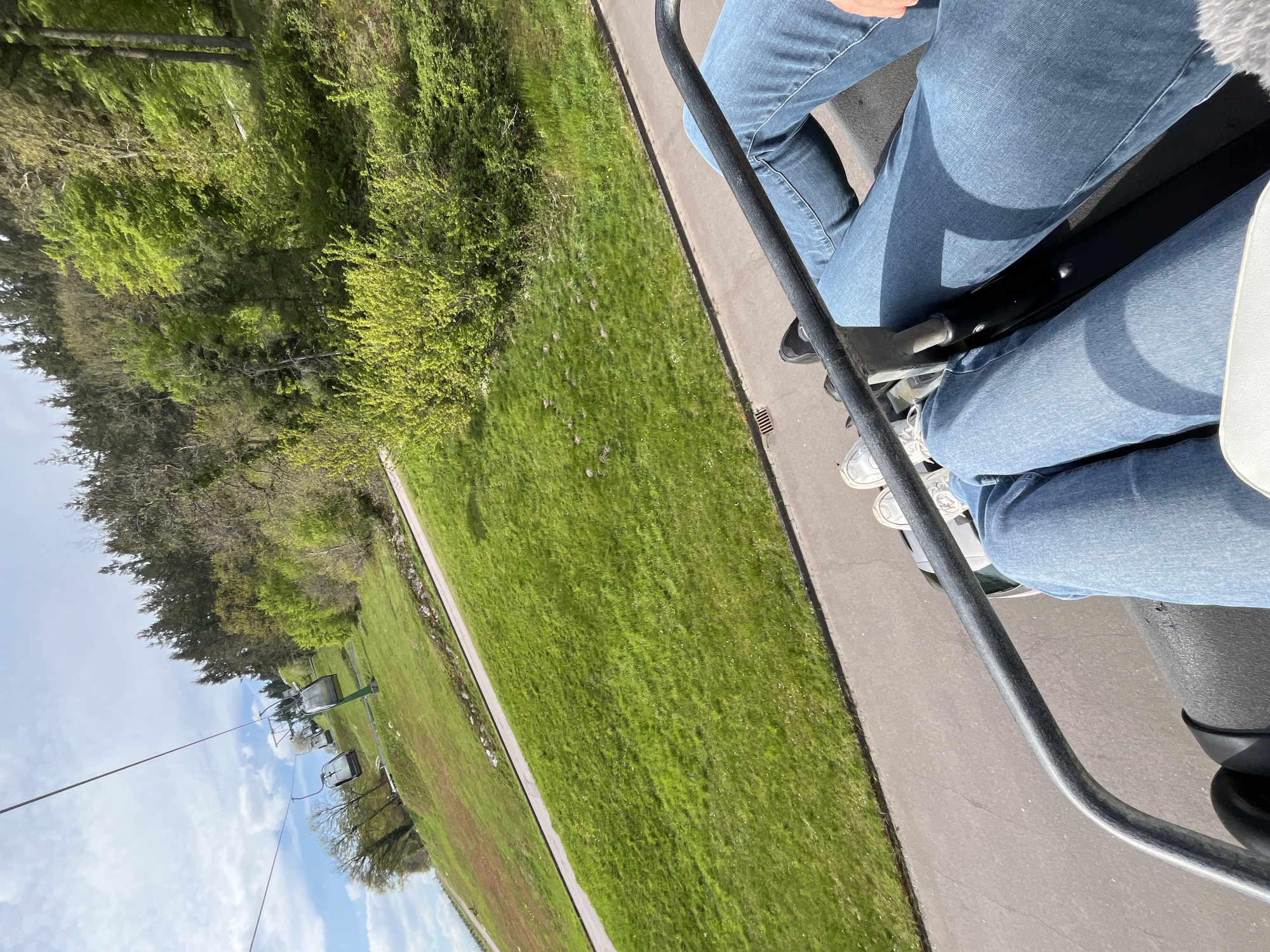 From the vantage point of a chairlift, two individuals are visible, both wearing blue denim jeans. The chairlift mechanism, including the safety bar, is in clear view. Below, a verdant lawn bordered by a paved path stretches out. The background features trees and a glimpse of other chairlifts. The location is Saarburg, Germany.