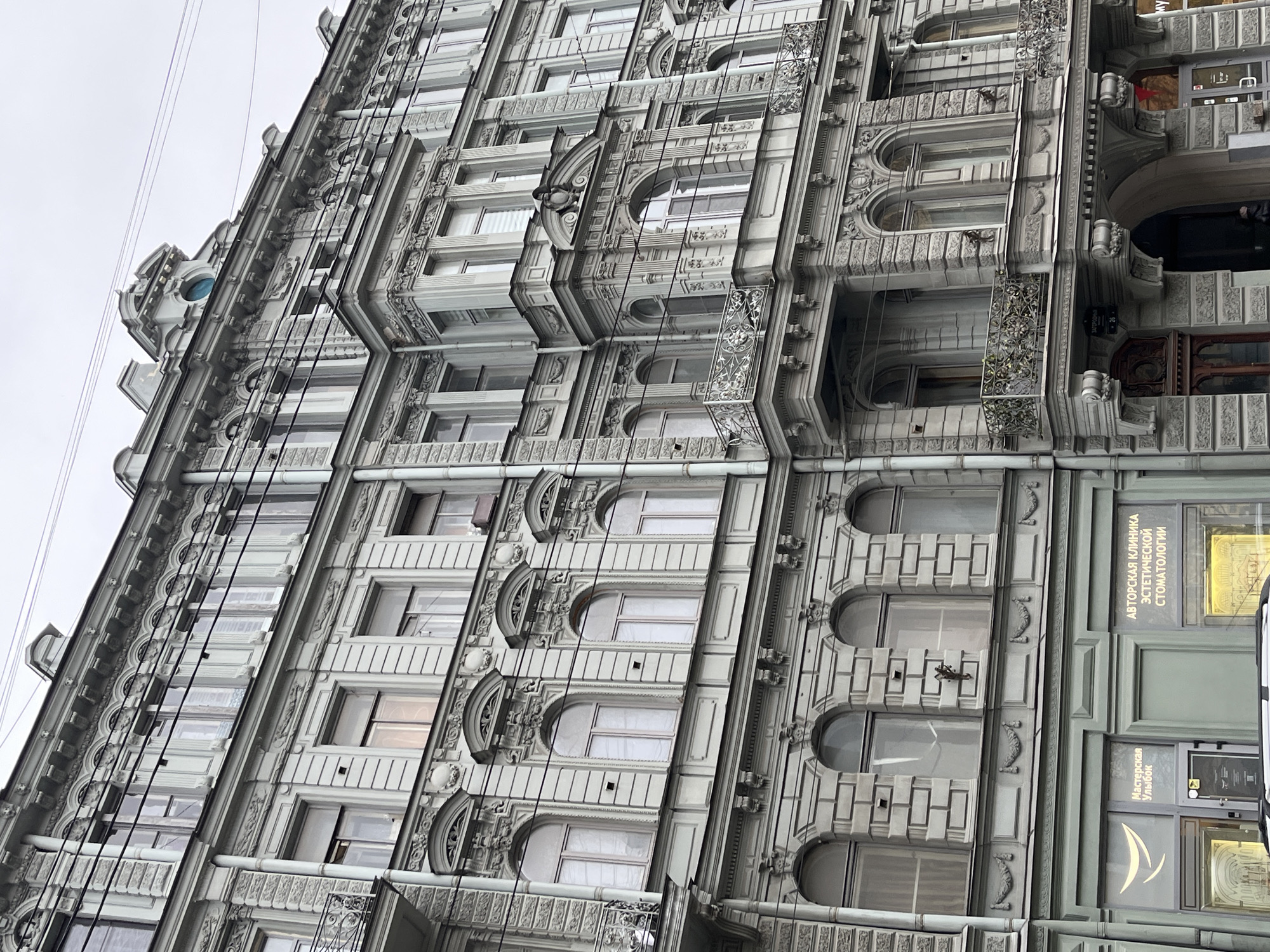 An upward-angled view captures the ornate facade of a multi-story building under an overcast, light gray sky. The building, in shades of light gray and off-white, exhibits a highly decorative architectural style characterized by intricate mouldings, cornices, pilasters, and various window designs.
The facade is rich in detail, featuring numerous windows that are rectangular, arched, or oval-shaped, many framed by elaborate decorative elements. Small, wrought-iron balconies or railings adorn some of the windows. The building's surface is heavily textured with classical and possibly Art Nouveau-inspired embellishments, including floral motifs and geometric patterns.
Several dark wires, likely utility lines, run vertically across the facade, partially obscuring the architectural details. The diffused light suggests an overcast or cloudy day.
On the lower right portion of the building, several signs with Cyrillic text are visible. One sign prominently displays "АВТОРСКАЯ КЛИНИКА ЭСТЕТИЧЕСКОЙ СТОМАТОЛОГИИ" (Author's Clinic of Esthetic Dentistry), and another below it reads "Мастерская Улыбок" (Workshop of Smiles). A small number "26" is also visible, likely an address. The architectural style combined with the Cyrillic text strongly suggests a location in a Russian city, such as St. Petersburg or Moscow.