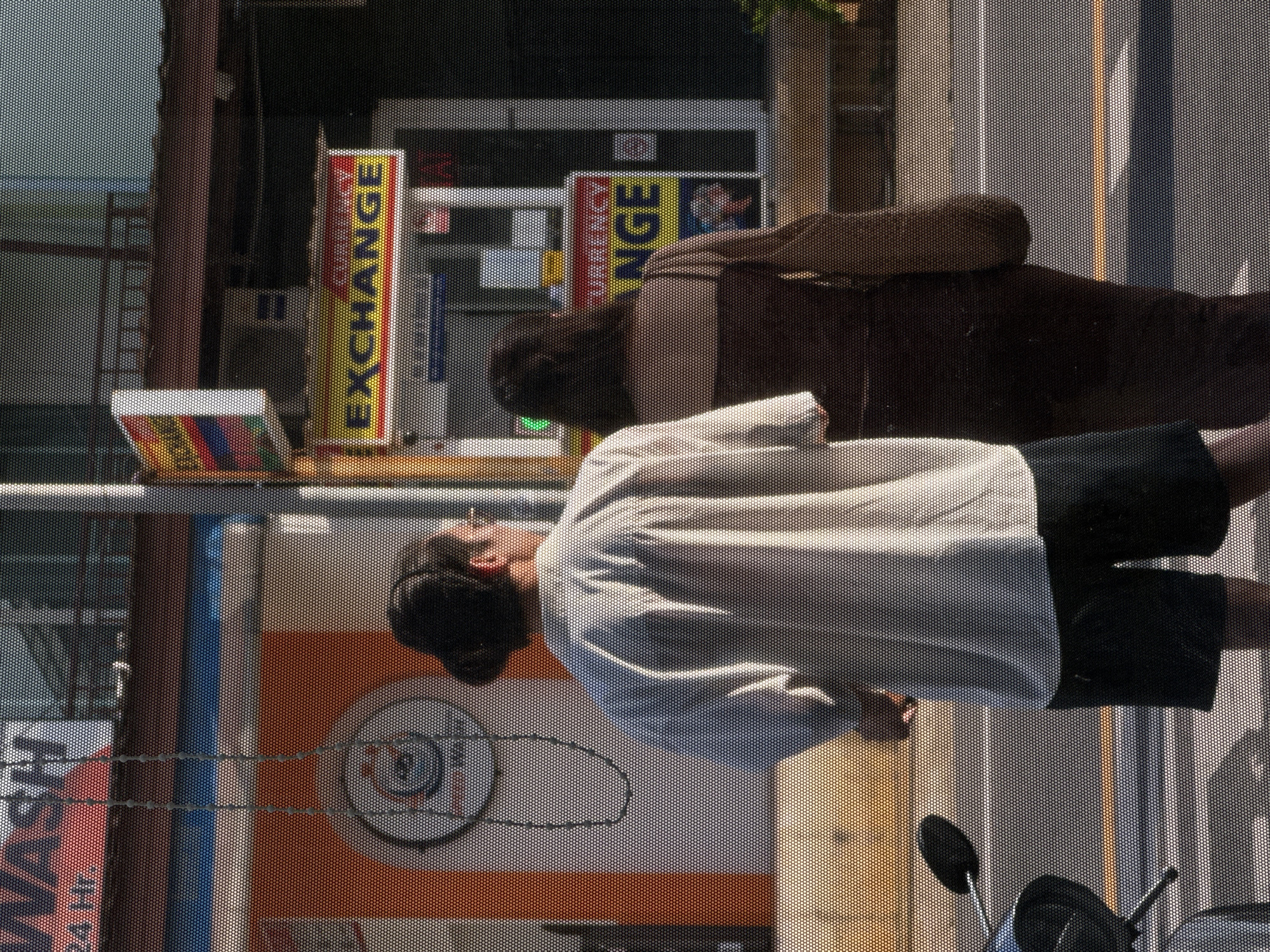 In Karon, Thailand, two individuals stand on a street with a currency exchange booth visible in the background. A man, facing away from the camera with his back to the viewer, wears a loose-fitting light-colored button-down shirt and dark shorts. His hair is pulled back. Beside him, a woman also faces away, dressed in a dark brown, form-fitting dress. To the left of the man, a portion of a motorcycle is visible, with its handlebars and front wheel showing. The street appears to be paved, with a yellow line marking the center. Behind the individuals, a building with a "WASH" sign is present, and further back, a brightly colored sign for "CURRENCY EXCHANGE" is prominently displayed. The scene is captured through a mesh screen, creating a textured effect on the image.