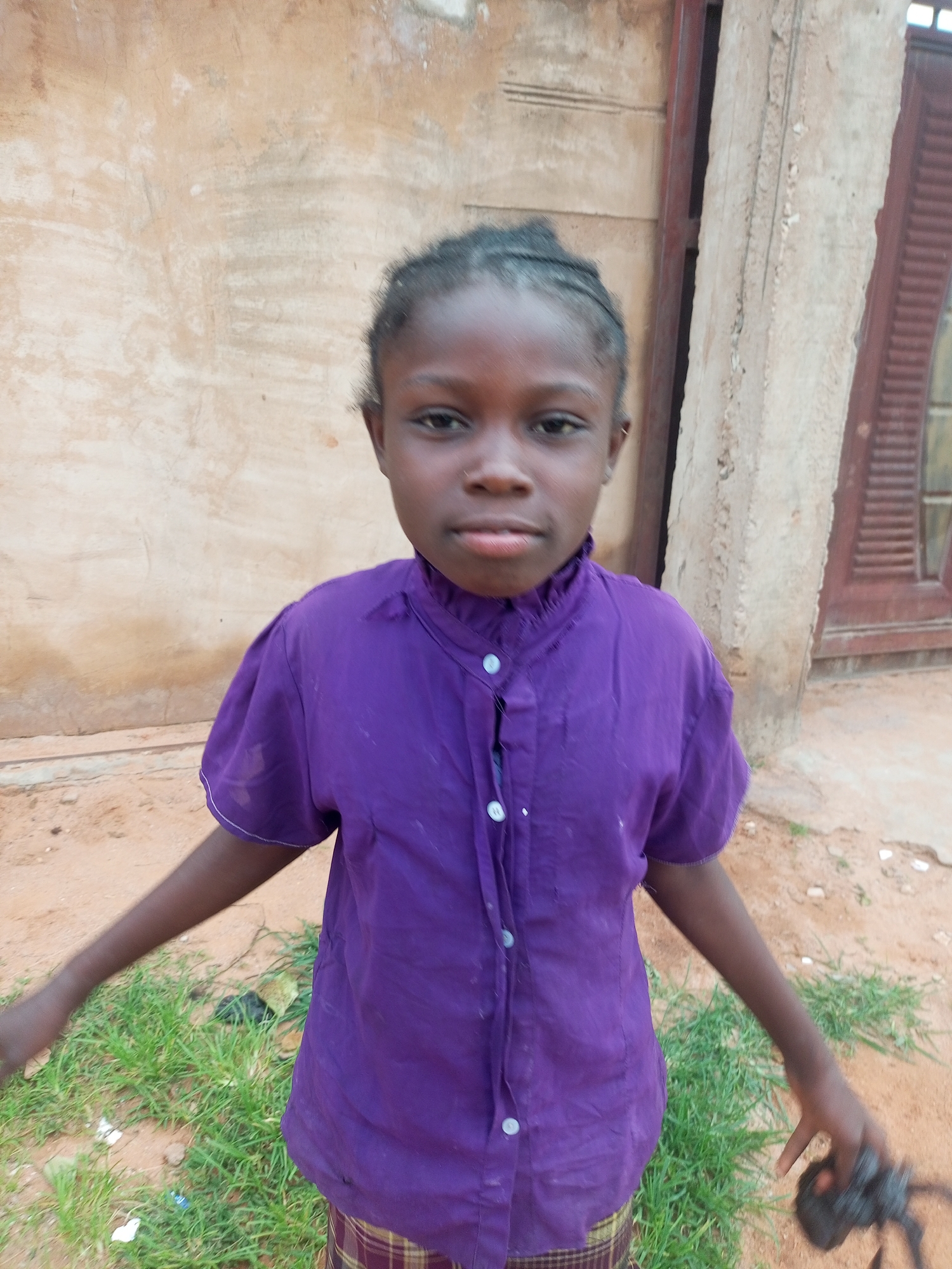 A young girl with dark skin and braided hair is standing in front of a textured, light brown wall. She is wearing a short-sleeved, collared purple shirt with buttons down the front and a plaid skirt. Her left arm is extended slightly outwards, and her right hand is holding a dark object, possibly a bird. The ground in front of her is a mix of sand and patches of green grass. To her right, there is a concrete pillar and a portion of a building with a dark brown door and slatted window. The overall scene appears to be outdoors in an urban or semi-urban environment, likely in Tempure, Nigeria.