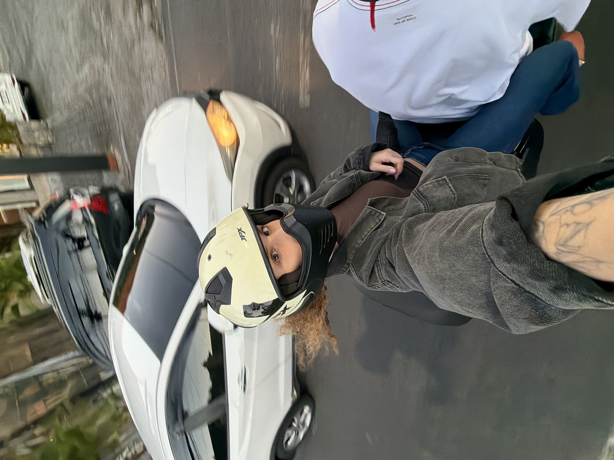 A first-person perspective selfie captures two individuals on a motorcycle, navigating a street in North Kuta, Indonesia. The rider, wearing a light yellow helmet with a black face mask, looks directly at the camera. Their curly brown hair is visible beneath the helmet. They are dressed in a dark denim jacket over a brown top and blue jeans. The passenger, in a white long-sleeved shirt, is partially visible to the right. The motorcycle is in motion, evidenced by the blurred background. A white car with its headlights on is to the left, and a dark-colored SUV is behind it. The environment appears to be an urban setting with paved roads and some greenery in the distance. The lighting suggests it is either early morning or late afternoon.