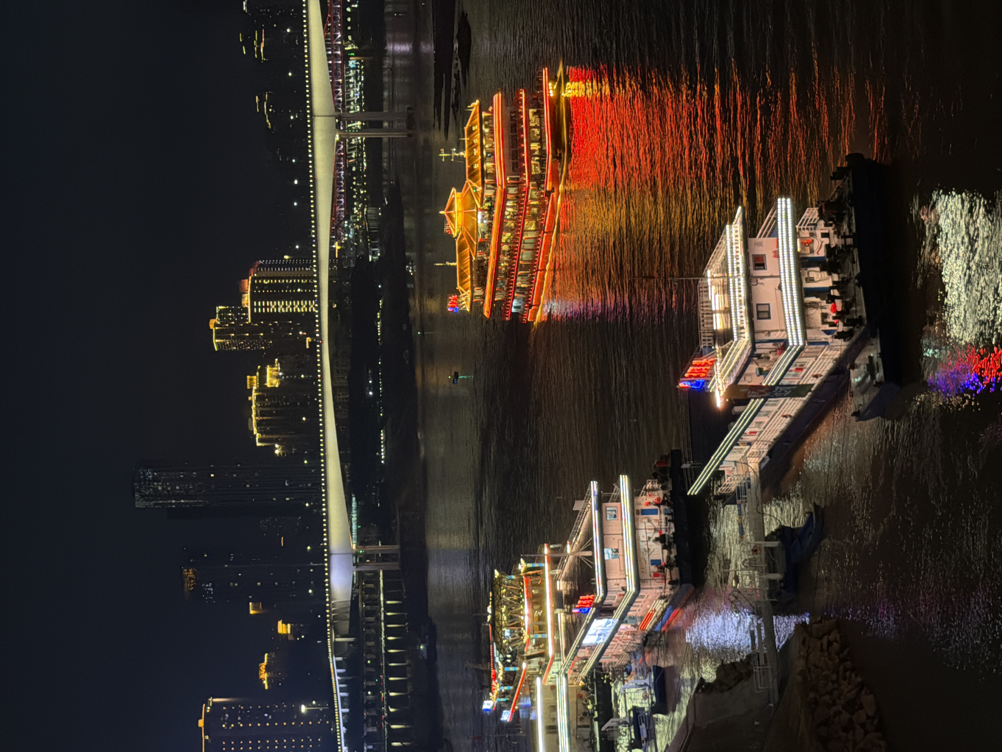 This media file depicts a nighttime scene of a vibrant cityscape along a river, likely in Yuzhong District, China. The primary subjects are illuminated buildings and a river cruise ship.
The scene showcases a modern cityscape with numerous tall buildings aglow with yellow and white lights, reflecting on the dark, rippling water. A prominent element is a large, multi-deck river cruise ship adorned with bright red and yellow lights that create a striking reflection on the water's surface. The ship appears to be docked or moving slowly. Another smaller vessel, also illuminated, is visible closer to the shore.
A long, modern bridge with a white superstructure and a railing lined with lights spans across the scene, connecting parts of the city. Parts of the bridge's lighting are visible, adding to the overall illuminated atmosphere. The water's surface is disturbed, with reflections of the lights creating a dynamic and colorful display.
The time of day is clearly night, indicated by the darkness of the sky and the extensive artificial lighting. The weather appears to be clear, allowing for good visibility of the city lights and reflections. The presence of the river and the dense urban development suggest a bustling port city.
There is no visible text in the media file. The overall mood conveyed is one of urban dynamism and nighttime beauty, highlighted by the interplay of lights and their reflections.