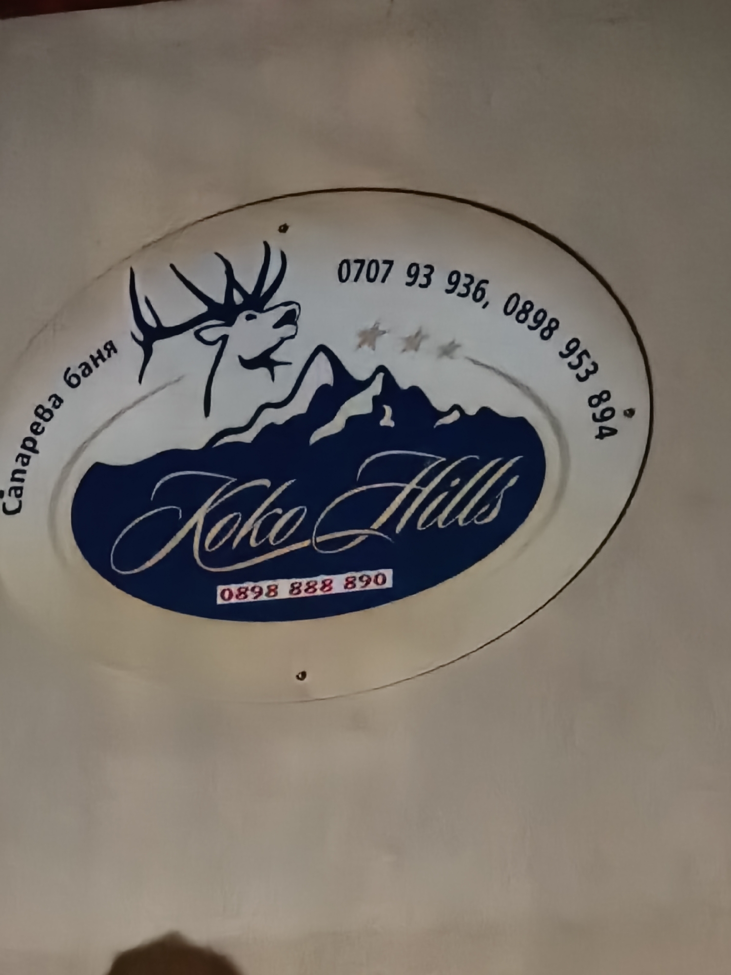 The image shows a sign for "Koko Hills" located in Сапарева баня. The sign is round and features a drawing of a deer and mountains. The phone numbers "0707 93 936, 0898 953 894" and "0898 888 890" are also displayed on the sign. The sign is white with dark blue lettering and graphics.