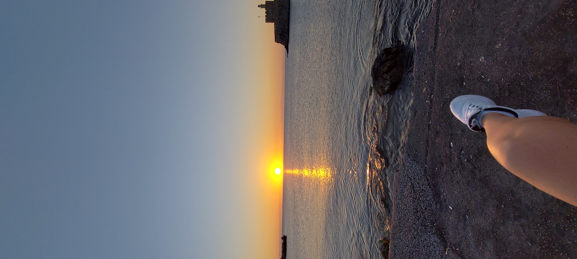 A low-angle, vertical shot captures a sunset scene by the sea in the Municipal Unit of Rhodes, Greece. The sun, a bright orb, sits on the horizon, casting a golden reflection across the calm water. In the distance, a medieval-style fortress with a lighthouse stands on a rocky outcropping. The foreground shows a person's lower leg and foot, clad in a white sneaker, resting on a dark, pebbly shore. The water laps gently at the shore, revealing submerged rocks. The sky transitions from a clear, pale blue at the top to soft oranges and yellows near the horizon.