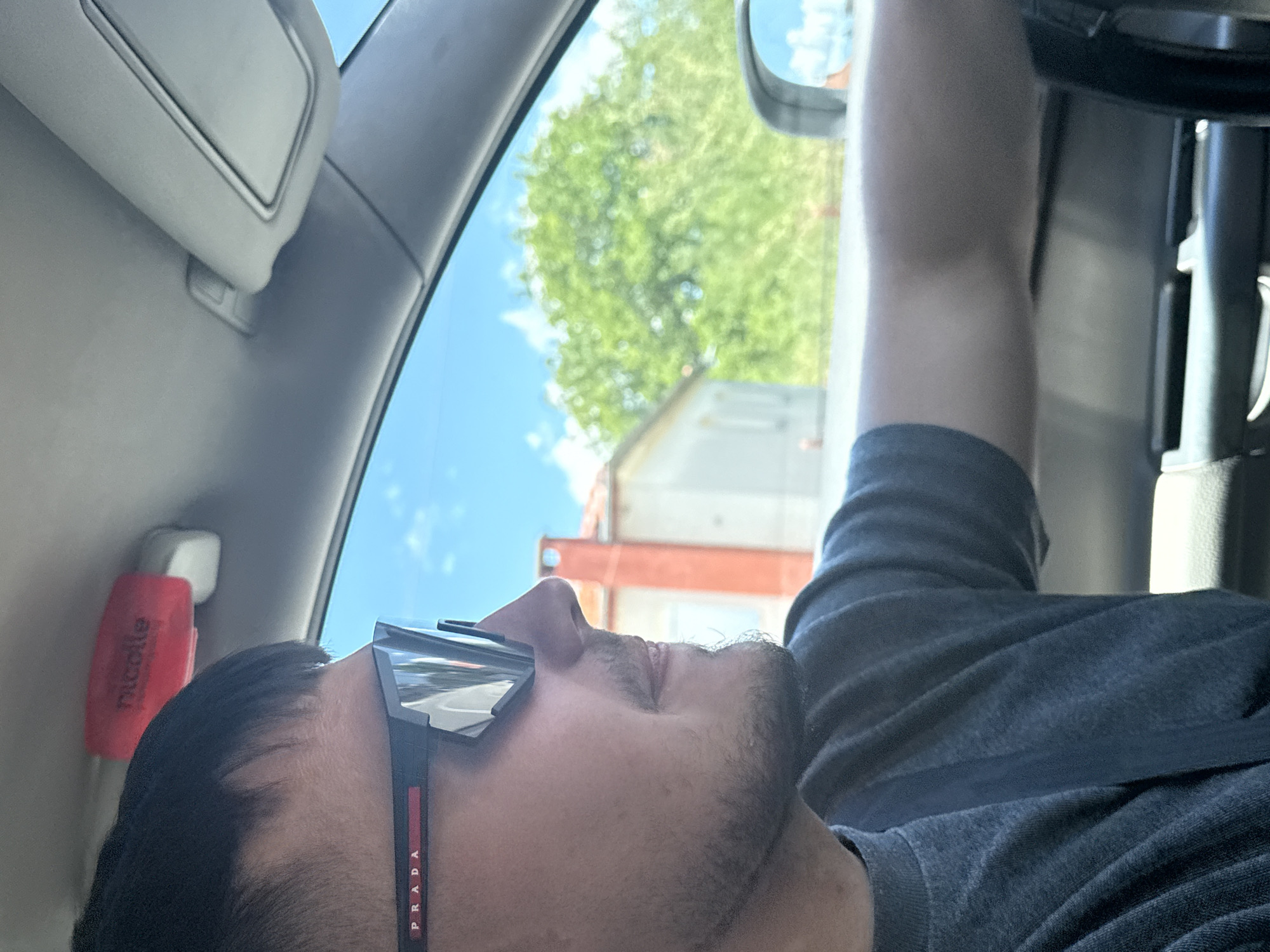 A man with short black hair and a goatee is seen from the side, driving a car. He is wearing black Prada sunglasses with a red stripe and a dark grey t-shirt. The interior of the car is visible, including the sun visor and a small red object with white text attached to the headliner. The view outside the car window shows a bright blue sky with some clouds, a house with an orange chimney, and lush green trees. A side-view mirror reflects the sky. The image appears to have been taken on a sunny day in Kato Nevrokopi, Greece.