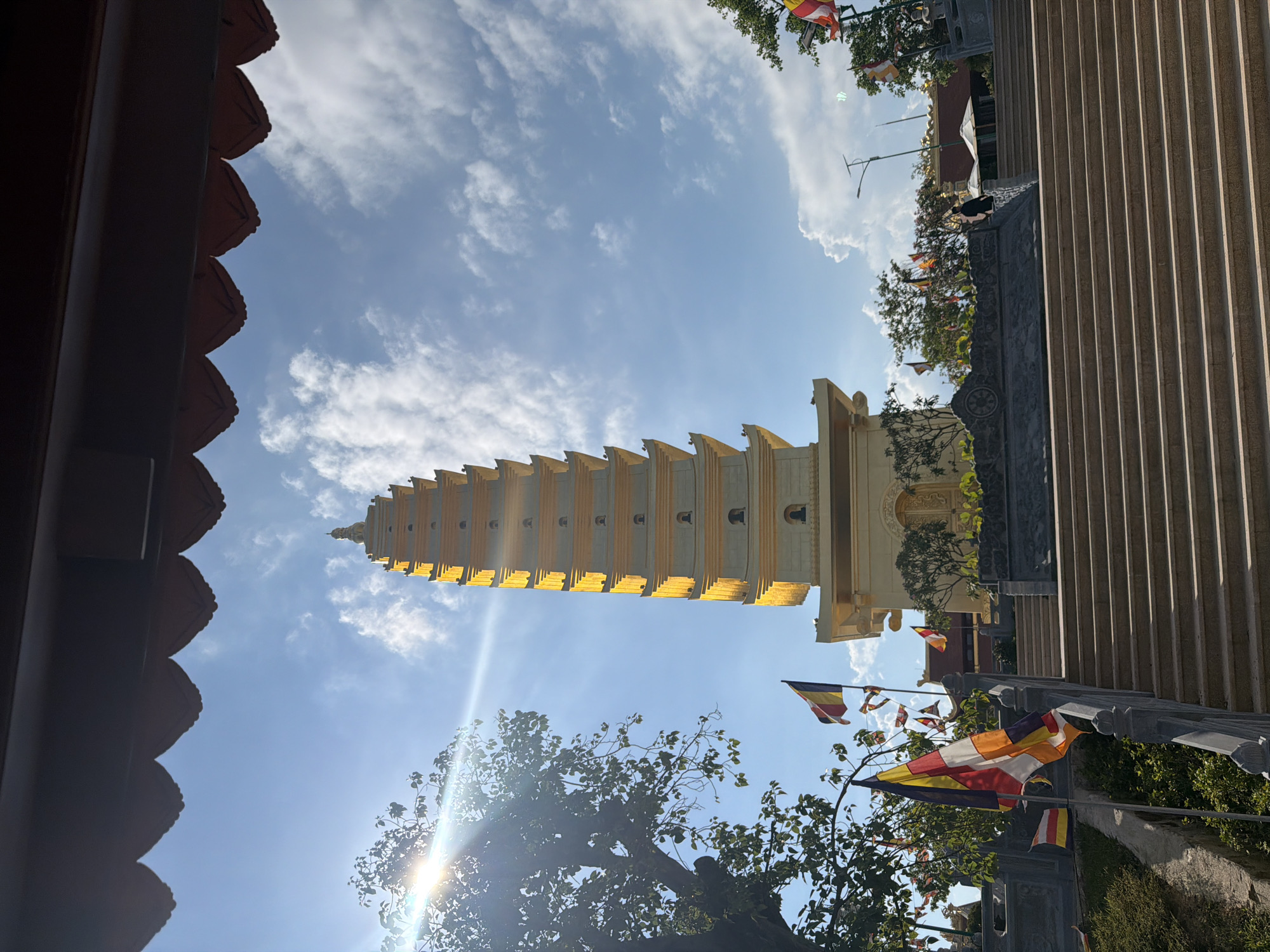 The image shows a multi-tiered pagoda in Nam Nha Trang Ward, Vietnam, viewed from a low angle. The pagoda is primarily light in color with golden accents. It stands against a partly cloudy blue sky. Buddhist prayer flags are visible. The lower portion of the image includes foliage and parts of the structure from which the image was taken.