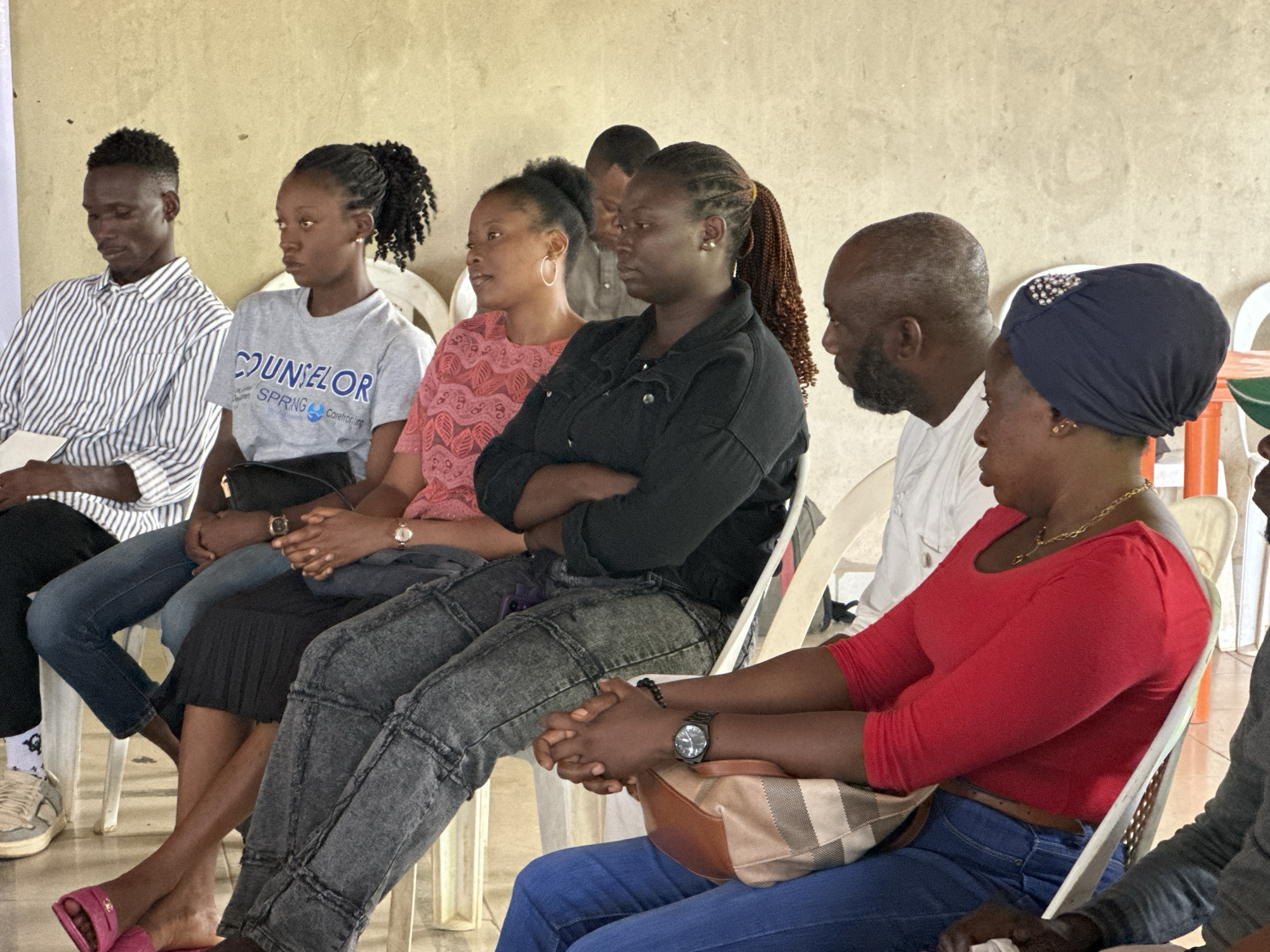 A group of approximately eight individuals, appearing to be adults, are seated closely together in chairs, facing mostly to the right of the frame, inside what appears to be a community hall or meeting space in Sabon Tasha, Nigeria. The walls of the room are a textured, light beige color, and the floor is not clearly visible.
In the foreground, a person wearing a bright red top and blue jeans is seated, with their hands clasped and resting on a beige and brown striped bag. Next to them, to their left, is a person wearing a black denim jacket and dark grey jeans, also with their arms crossed. Another individual in a pink patterned top is seated to the left of the person in the black jacket.
Further to the left, a person in a grey t-shirt with "COUNSELOR" and "SPRING Carefronting" printed on it is seated, their arms resting in their lap. Beside them, a person in a black and white striped shirt is visible. Partially obscured in the background are other individuals, including a man with a bald head and beard, wearing a white shirt.
The lighting in the scene appears to be natural, coming from an unseen source, creating soft shadows. The overall impression is that of a gathering or a meeting in progress.