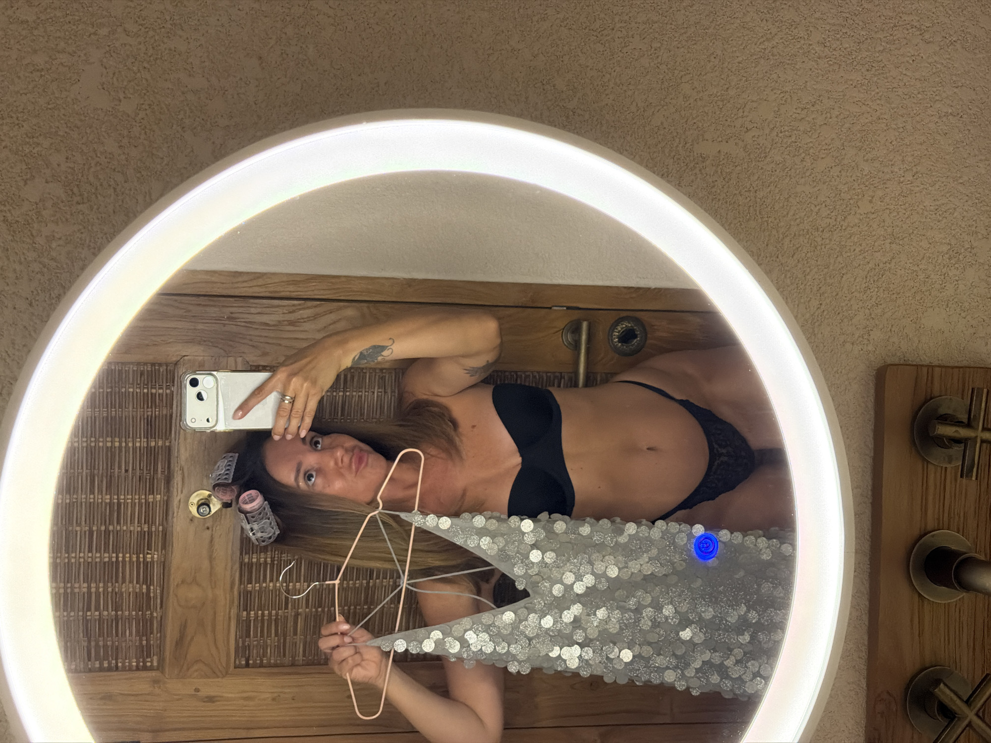A woman is taking a mirror selfie in a bathroom. She is wearing a black bra and black thong. In her right hand, she is holding a hanger with a sequined top. She has hair rollers in her hair. The bathroom has wooden walls and a textured wall on the right. A circular mirror with an LED light is visible. The image is taken in a bathroom located in Mengwi, Indonesia.