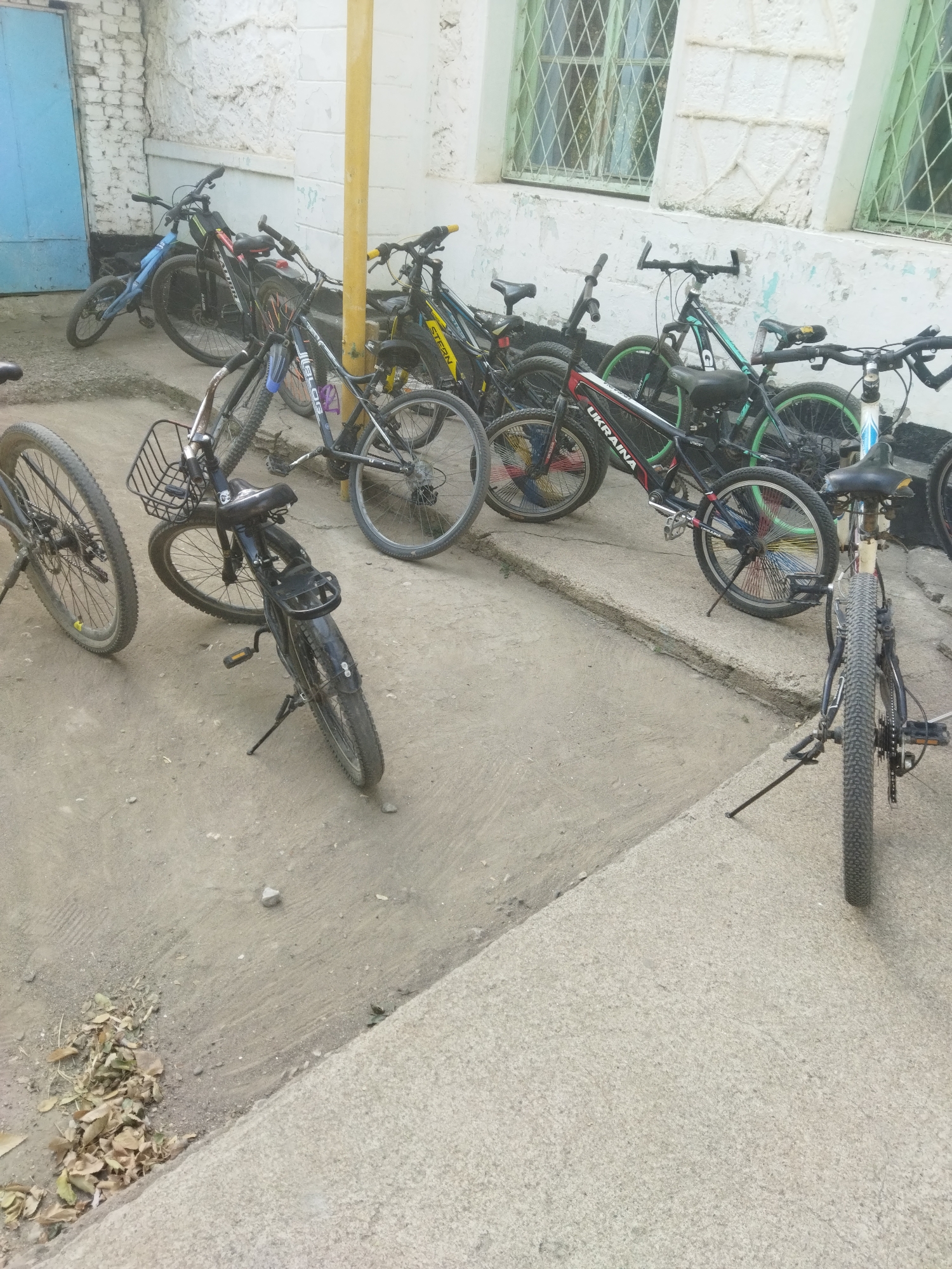 The image depicts an outdoor scene with multiple bicycles parked on a concrete and dirt surface adjacent to a building with a white, textured wall and green-framed windows. On the left, a blue bicycle with a small frame is parked near a light blue door. Several other bicycles are visible, including one with a basket on the handlebars and a black frame with "UKRAINA" branding. Another bicycle has colorful spokes. The bicycles appear to be of various types and conditions. The environment suggests an urban or semi-urban setting.