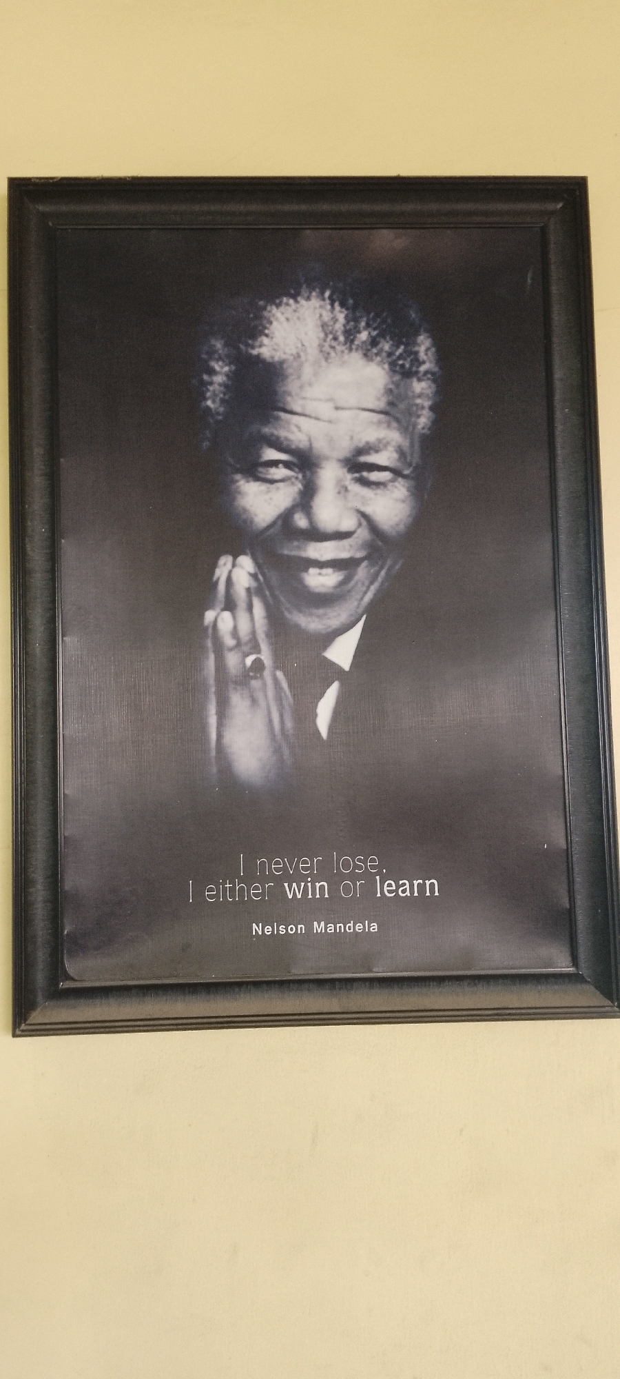 A framed black and white portrait of Nelson Mandela hangs on a light yellow wall. The portrait depicts Mandela smiling with his hands clasped together. Below the image, the text reads "I never lose, I either win or learn," followed by "Nelson Mandela." The frame is dark colored, and has a slight sheen. The image is taken at eye level. I do not have the ability to retrieve the location in the city of Akwanga, Nigeria.