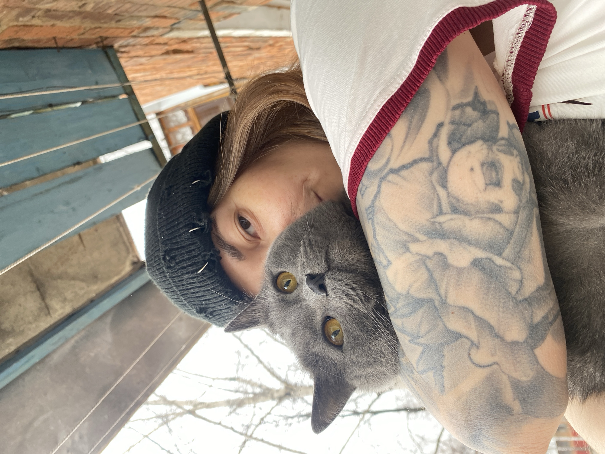 A person with light brown hair and a black beanie is holding a gray cat. The person is wearing a white shirt with a red trim. The person's left arm is visible and has a large floral tattoo. The background shows a partly visible, weathered blue wooden structure and bare tree branches. The photograph appears to have been taken outdoors on a day with overcast light.
Although the prompt mentioned Votkinsk, Russia, the image itself does not contain explicit visual information confirming the specific location. Therefore, the description does not include this detail.