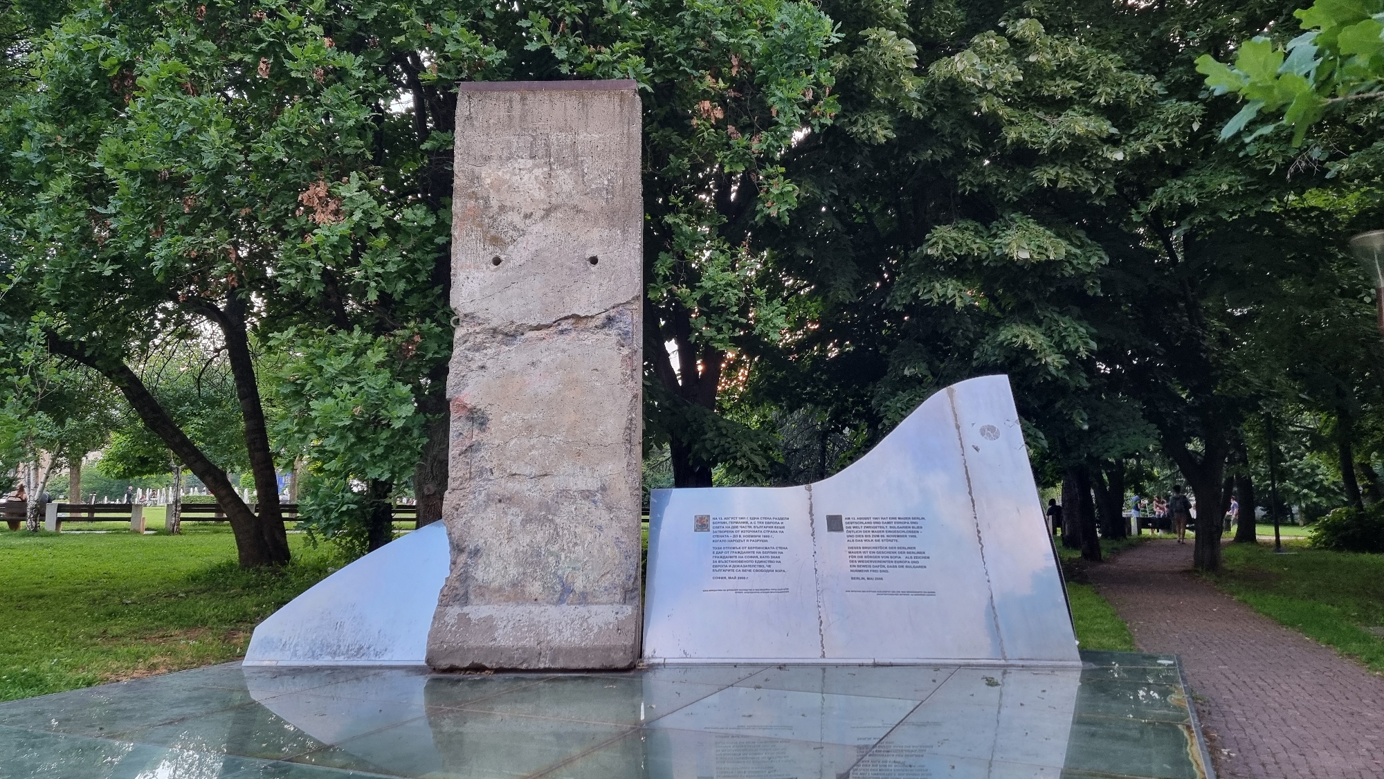 This media file captures a memorial in an outdoor park setting, likely in Sofia, Bulgaria, as indicated by the location context. The central subject is a fragment of the Berlin Wall – a weathered concrete slab with visible cracks and two circular holes, standing upright. To its right, a curved metallic plaque with text in both Bulgarian and German serves as an explanatory memorial. Both the concrete piece and the plaque rest on a reflective, possibly glass, base which shows distorted reflections of the structure and the surrounding greenery.
The scene is set in a lush green park with numerous trees, some featuring large, leafy canopies, and a grass lawn. In the background, benches and a dirt path are visible, along with a few indistinct figures walking further down the path, suggesting a public area. The lighting appears soft, indicative of daytime, possibly late afternoon or early evening, with some sunlight filtering through the dense foliage.
The visible text on the plaque, in Bulgarian on the left and German on the right, commemorates the historical significance of the Berlin Wall. It states that on August 13, 1961, a wall divided Berlin, Germany, Europe, and the world. It further notes that from November 9, 1989, to November 20, 1990, the wall was peacefully dismantled. The fragment is described as a gift from the citizens of Berlin to the citizens of Sofia, symbolizing a reunited Europe and serving as proof that Bulgarians were among the winners of this historical change. The plaque also bears the dates "Sofia, May 2006" and "Berlin, May 2006".
