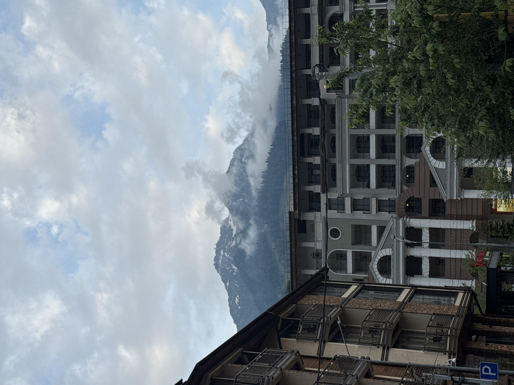 The image depicts a view of mountainous terrain partially obscured by clouds, with several buildings in the foreground. The architecture of the buildings appears to be modern, featuring multiple stories, balconies, and decorative columns. A prominent mountain range, with snow-capped peaks and forested slopes, rises in the background. The sky is cloudy, with patches of blue visible. The scene suggests a resort or urban area located in a mountainous region, likely Estosadok, Russia, known for its natural beauty and recreational facilities. No people are visible in the image.