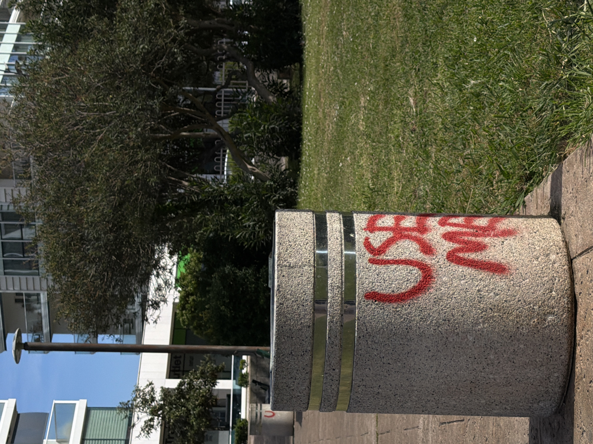 The image depicts an outdoor urban scene, rotated 90 degrees clockwise from its natural orientation. If viewed upright, a large, cylindrical concrete bollard dominates the right foreground, resting on a concrete path. The bollard is made of speckled concrete aggregate and features two horizontal, shiny metallic bands near its top. Bold red spray paint, forming illegible graffiti that appears to spell "SUSH" or a similar stylized word, is scrawled across the top and side of the bollard.
To the left of the bollard and extending into the midground is a vibrant green grassy area, which looks well-maintained with some lighter patches and small white wildflowers scattered within. Beyond the grass, a variety of trees and bushes with dense green foliage create a natural barrier.
In the background and extending along the left side of the frame, modern multi-story buildings are visible. These buildings feature numerous windows, some with reflective surfaces, and balconies, suggesting an apartment or commercial complex. A tall, slender dark light pole with a flat, circular light fixture stands prominently in front of one of these buildings on the left. Partial text "tlet", likely part of "Outlet", is visible on a sign on one of the buildings. The scene is illuminated by bright sunlight, indicating a clear, sunny day in an urban park or promenade setting, consistent with the Budva, Montenegro context.