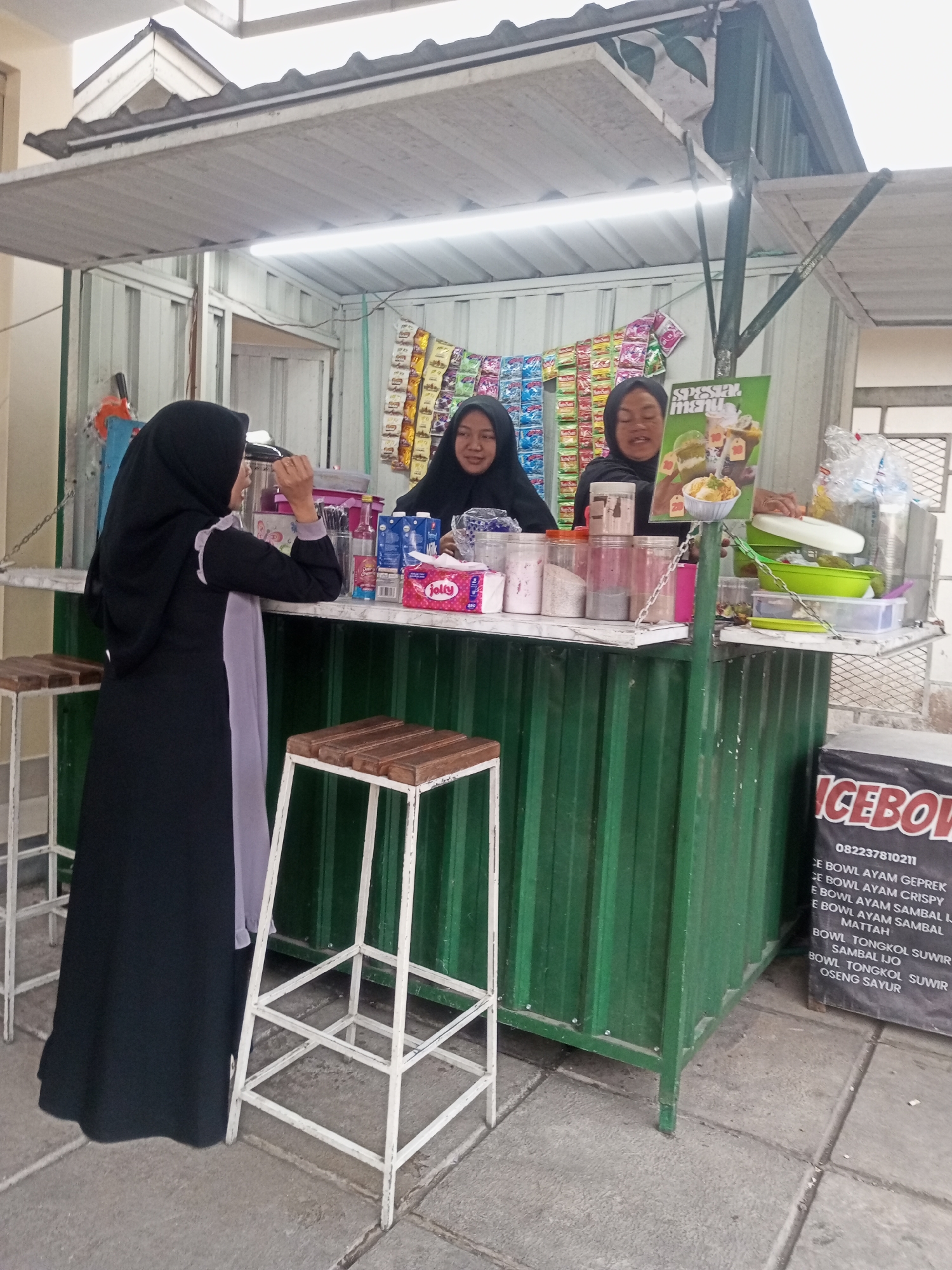 In Pandansari, Indonesia, two women are attending a food stall. One woman, wearing a black hijab and abaya, stands to the left of the stall, holding a bottle and looking at it. The second woman, also in a black hijab, stands behind the counter, facing the first woman and smiling. Behind the counter, shelves are stocked with various food items and beverages. A green metal structure forms the front of the stall, with a white counter above it. Two white stools with wooden seats are positioned in front of the counter. A sign with the text "ICEBO..." is visible on the right side of the image. The ground is paved with concrete tiles.