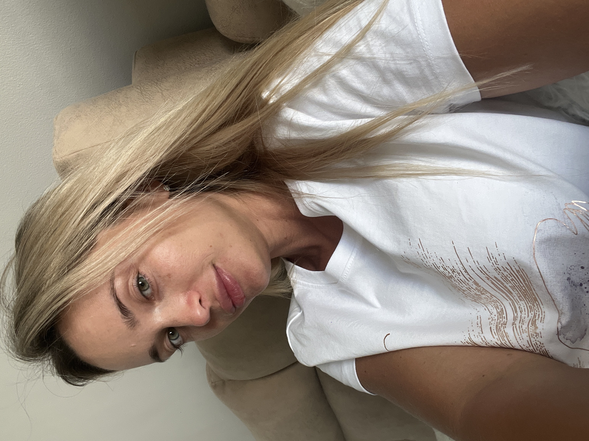A young Caucasian woman with blonde hair and green eyes is captured in a close-up, selfie-style shot. She is wearing a white t-shirt with a subtle rose gold abstract design. The background consists of a beige upholstered sofa and a light-colored wall, suggesting an indoor setting. The lighting appears natural and soft, possibly from a nearby window. No specific location in Sharm El Sheikh, Egypt, can be determined from the visual information provided.