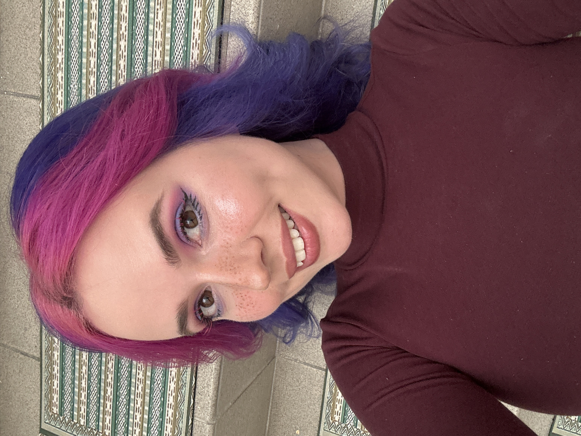 A sideways-oriented selfie captures a smiling young woman from the chest up. She gazes directly at the camera with a broad, joyful smile, revealing her white teeth. Her hair is strikingly two-toned: vibrant magenta on her right side and deep purple on her left side. She has light skin, noticeable freckles across her nose and cheeks, and brown eyes accentuated by prominent eyelashes and purple eyeshadow. She wears a high-necked, long-sleeved top in a solid dark maroon or burgundy color.
The background features a decorative patterned surface, possibly a mat, rug, or tiled wall/floor, displaying vertical stripes and geometric diamond motifs in shades of green, white, and beige. This is set against a textured, light grey stone or concrete surface. Given the location context of Zagreb, Croatia, this pattern may represent local design or architecture. The bright, natural lighting suggests the photo was taken during the day. No text is visible in the image.