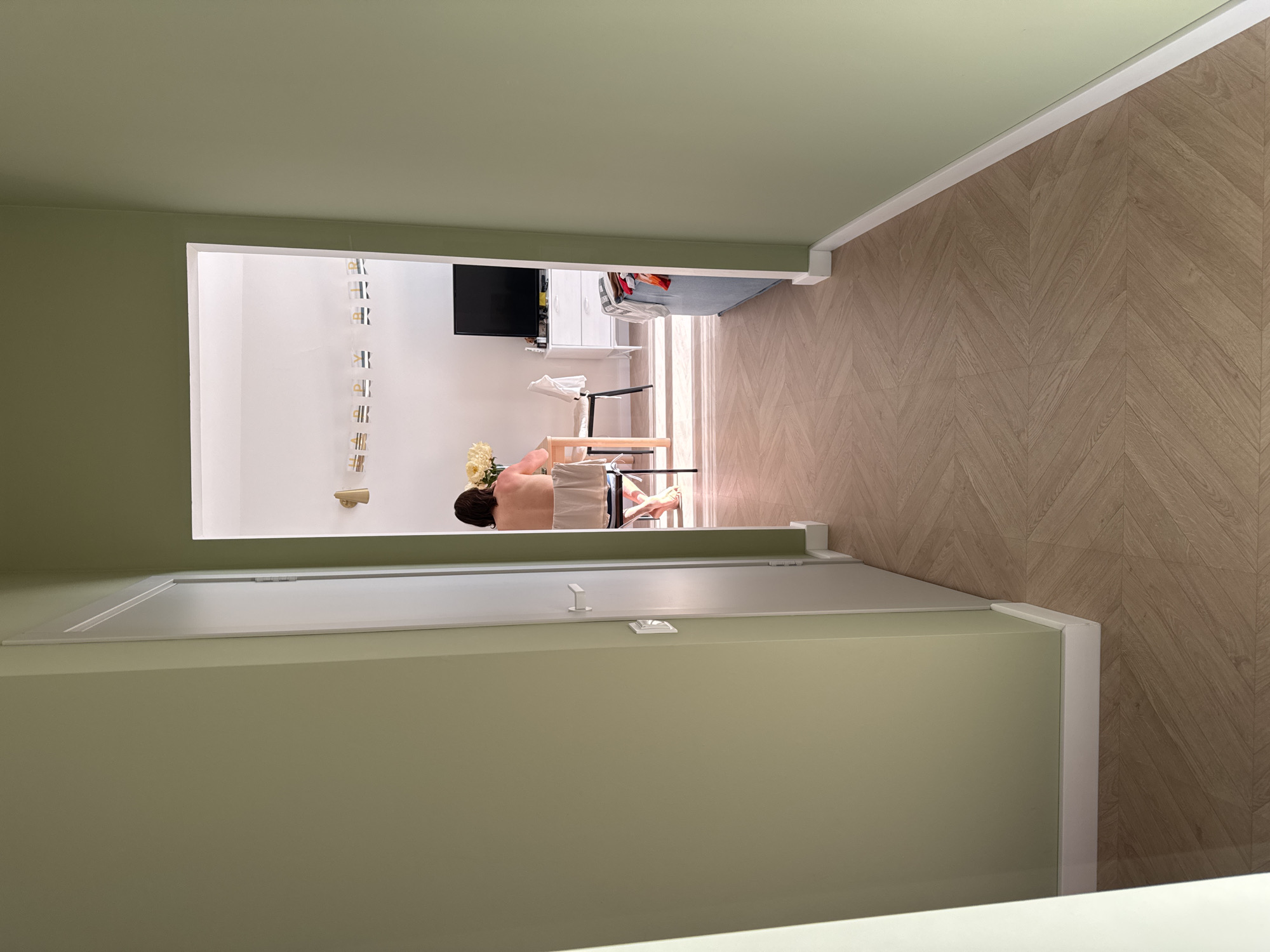 The image presents a view from an interior space, likely an entryway or hallway, looking into another room. The foreground features a doorframe, painted in a light green color. Beyond the doorway, the room has white walls and light wood-toned flooring with a chevron pattern. In the room, a person is seated at a small table or desk, and there are flowers on the table. A television is mounted on the wall in the background. The image is taken from an elevated perspective. It is not possible to determine the location in the city of Люк, Russia.