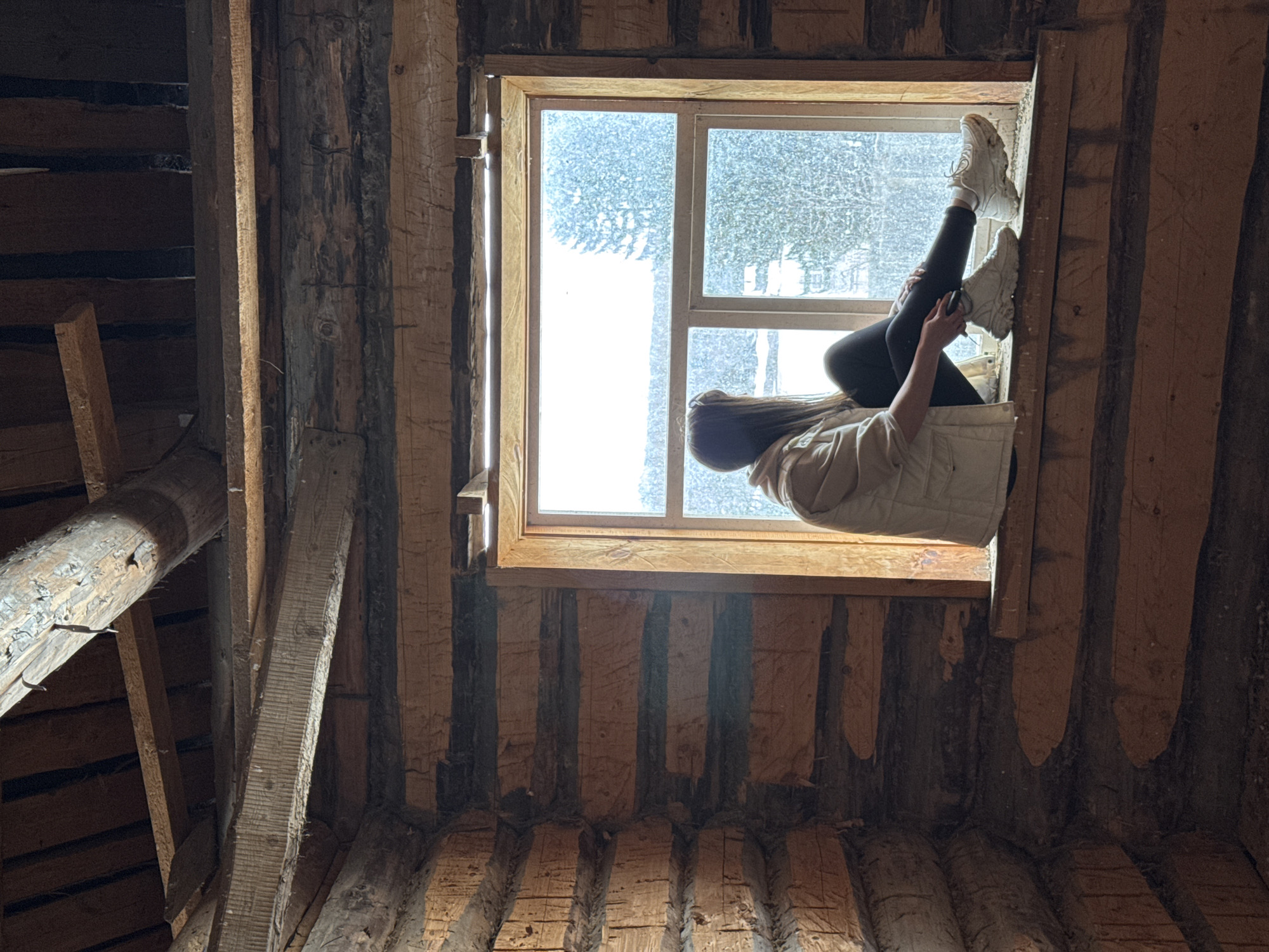 The image depicts a person sitting in a window frame within a rustic, wooden structure. The person is wearing leggings, white sneakers, and a light-colored jacket, and is positioned with their legs extended upwards and leaning against the window. The exterior view through the window is a blurred, light-colored landscape, possibly snowy. The structure's interior walls are constructed from rough-hewn logs with visible chinking, creating a cabin-like environment. The scene is illuminated by natural light filtering through the window. The city of Amaneevo, Russia, is mentioned as the location.