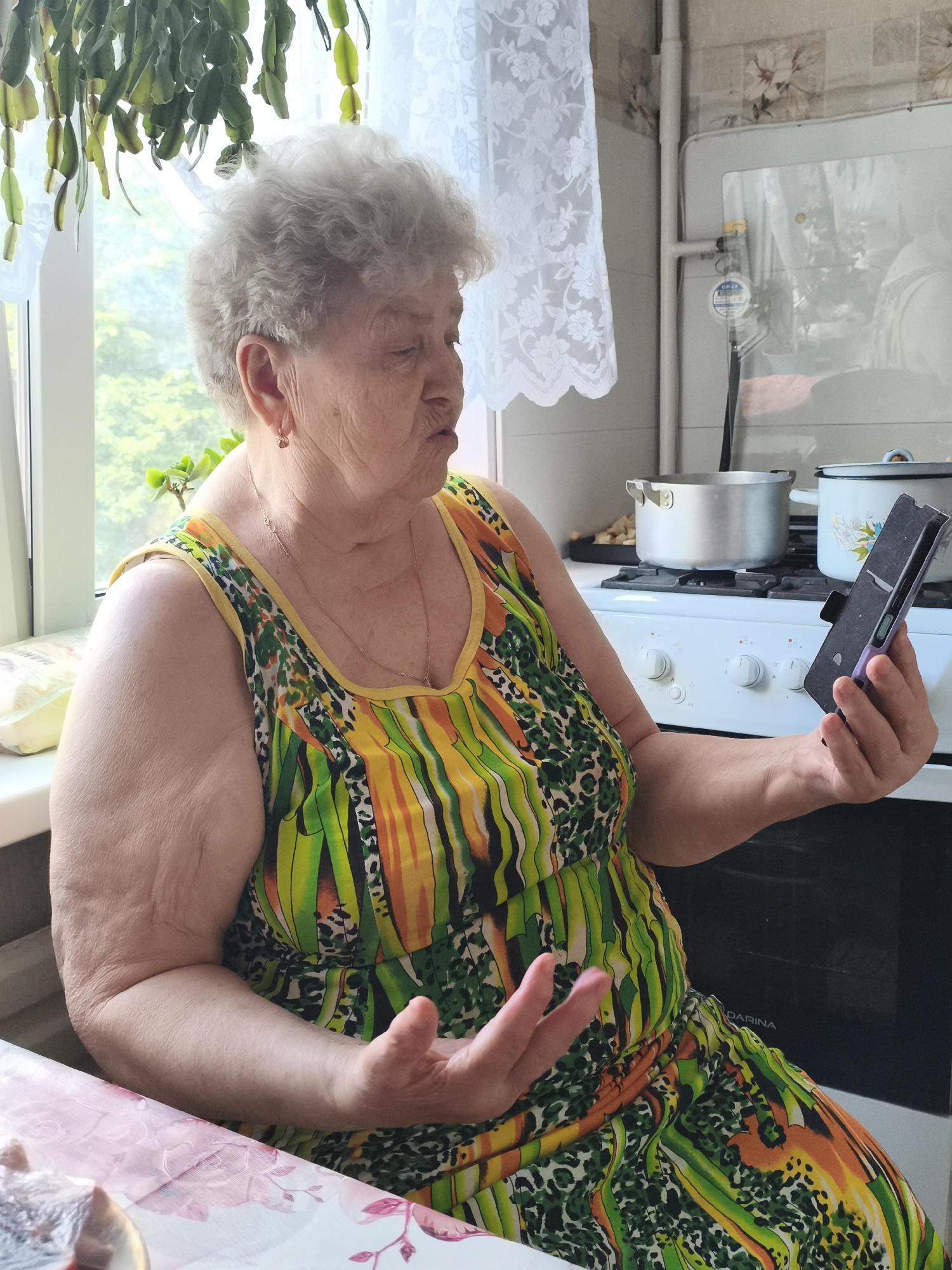 The image depicts an elderly woman in a brightly colored, sleeveless dress sitting at a table in a kitchen. She is holding a smartphone in her right hand and gesturing with her left hand. The kitchen appears to be in a residential setting, possibly an apartment, with a stove, pots, and a window visible in the background. There is a plant hanging above the window. The woman has gray hair and is looking at the phone. A floral-patterned tablecloth is visible on the table in the foreground. Based on the visible text "DARINA" on the stove, which is a brand of kitchen appliances common in Russia, and the overall aesthetic, the image is likely from Izhevsk, Russia.