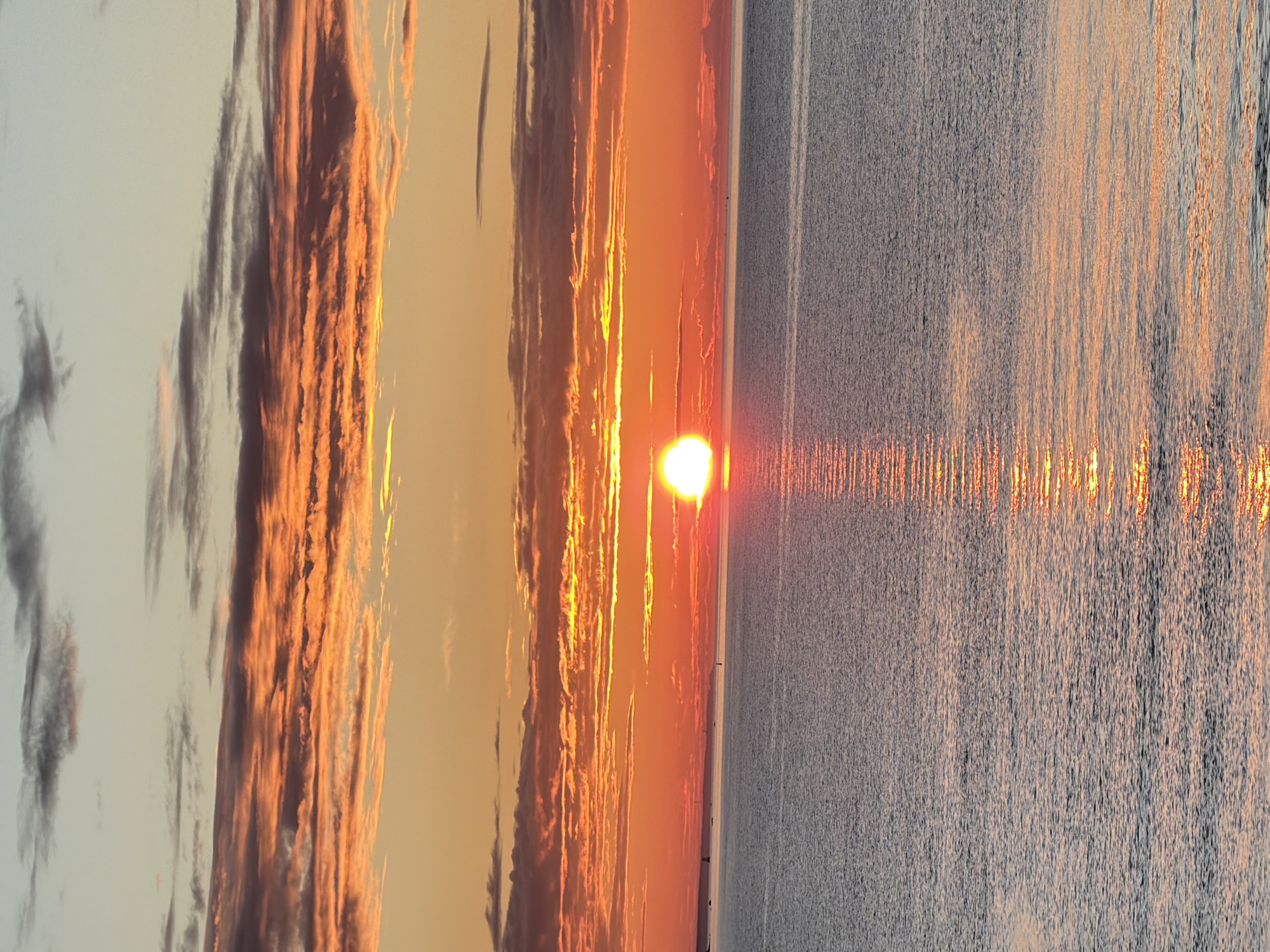 This is a photograph of a sunset over the Baltic Sea, viewed from Tallinn, Estonia. The sun, a bright white orb, is low on the horizon, casting a golden reflection on the water. The sky is ablaze with oranges, reds, and yellows, with streaks of dark clouds adding depth and contrast. The sea is calm, with gentle ripples reflecting the colors of the sky. The coastline is visible in the distance, with a few faint structures that could be lighthouses or buildings. There are no people or animals visible in the image. The overall impression is one of peacefulness and natural beauty.