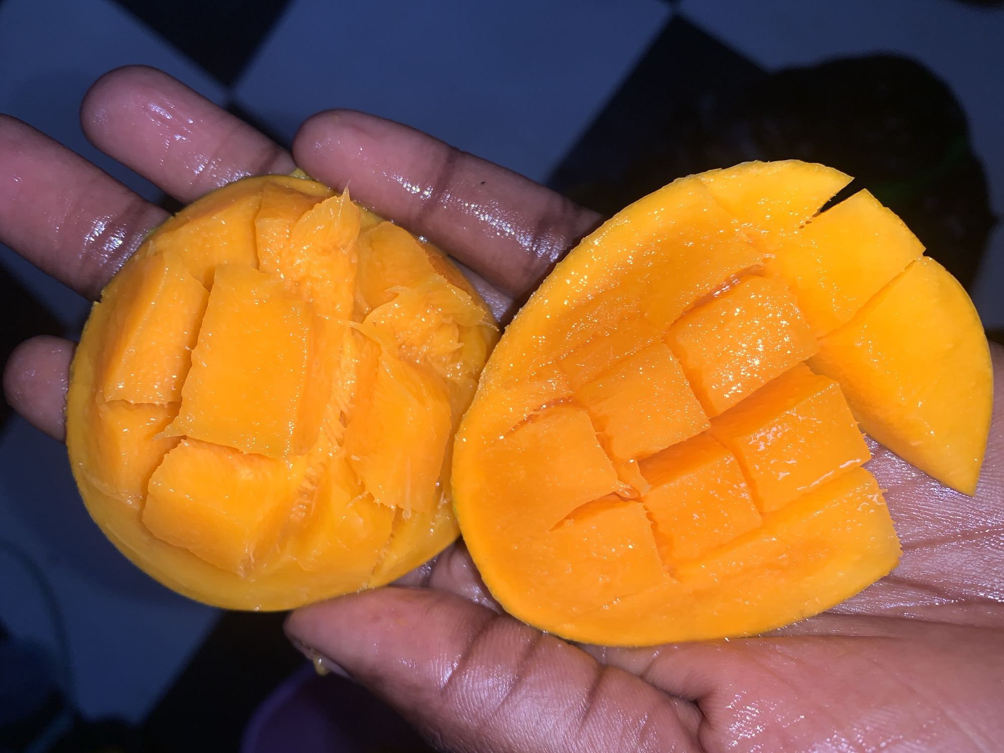 A close-up shot shows a pair of hands holding two halves of a mango. Each half of the mango has been scored into a grid pattern, with the fleshy cubes partially detached. The skin of the mango is not visible in the photograph. The hands appear to be slightly wet or oily. The background is blurry, showing a hint of a checkered pattern on what might be a surface.
The image does not contain enough information to confirm the location in the city of Rafin Par, Nigeria.
