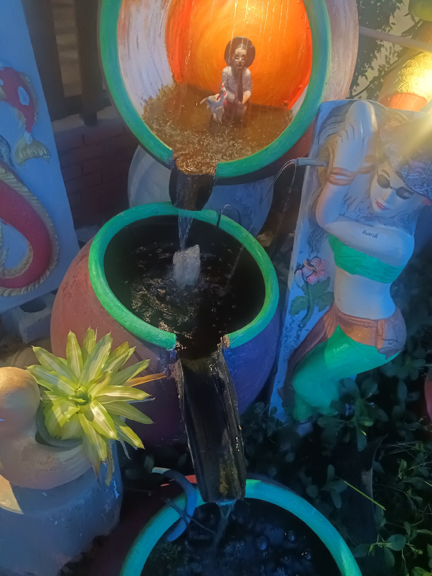 The image depicts a tiered water fountain structure located in Ban Nong Khon, Thailand. The fountain consists of four pots stacked vertically, each a different size and color (orange, purple, green, blue), all with a green rim. Water cascades from the top orange pot down into the pot below in a cascading pattern. There is a statue of an elderly woman, holding an animal, within the top orange pot. Next to the pots, there is a statue of a reclining woman in sunglasses, with Thai lettering on the structure behind the reclining woman. There are decorative panels with what appears to be a red and white fish on one. A potted plant is visible near the base of the structure. Dark foliage surrounds the base of the fountain, and a dark background suggesting night time.