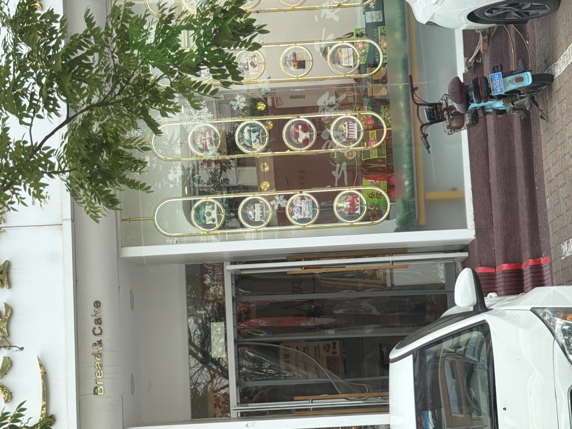 The image shows the exterior of a bakery named "Bread & Cake." The bakery's windows are decorated with golden circular frames containing images of cakes. Outside, a white vehicle is parked on the right side, and a parked blue scooter is near the entrance. The location appears to be in an urban area, likely in Guta, China, given the context.