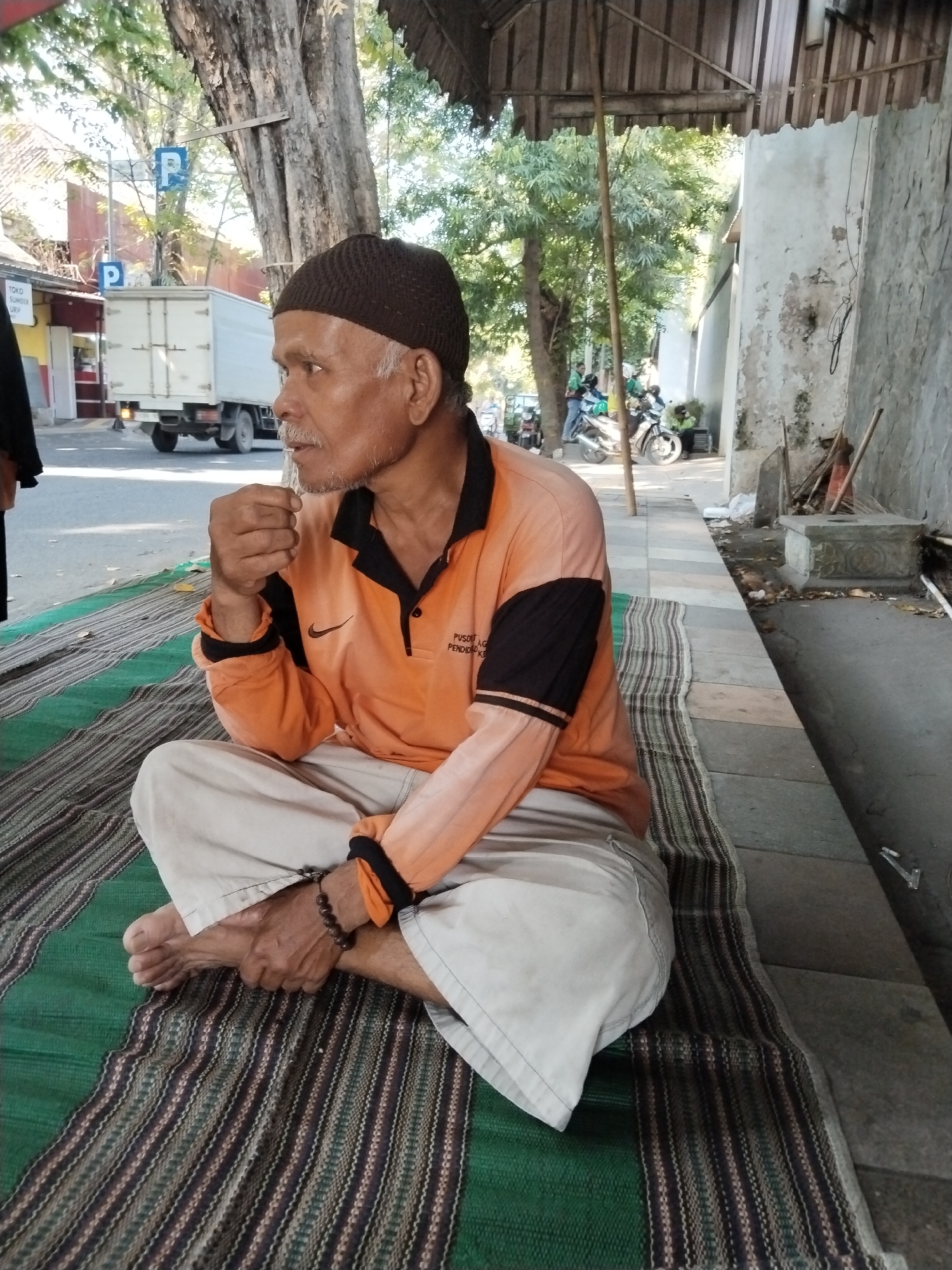 A man with a brown skullcap and a mustache is sitting cross-legged on a woven mat on a sidewalk. He is wearing an orange polo shirt with black accents on the sleeves and collar, and beige pants. He is holding something small to his mouth with his right hand. He has a beaded bracelet on his left wrist. The mat is placed on a paved area next to a textured wall. Behind him, across a street, is a white truck with a parking sign and trees. Further down the street, there are motorbikes and people. The scene appears to be in an urban environment, possibly along a road in Kecamatan Pati, Indonesia.