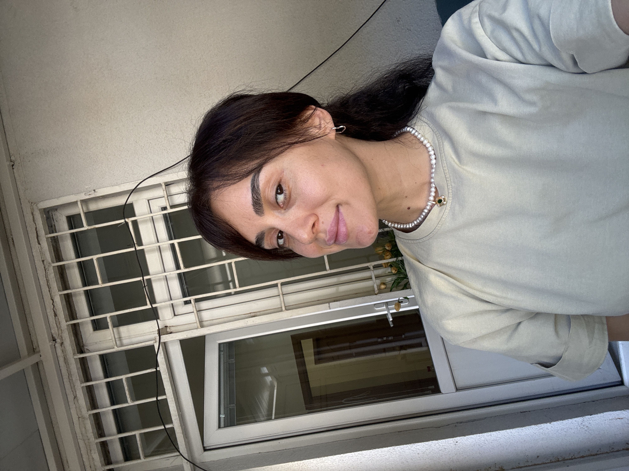 A woman is taking a selfie in front of a building with barred windows and a closed door. The woman has dark hair, is wearing a pearl necklace with a green pendant, and a light-colored t-shirt. She is looking at the camera with a slight smile. The background shows a building with white bars on the windows, a white door with a glass panel, and some yellow flowers on the right side. The overall setting appears to be an outdoor space, possibly a courtyard or balcony, in Tbilisi, Georgia. The lighting suggests it is daytime.