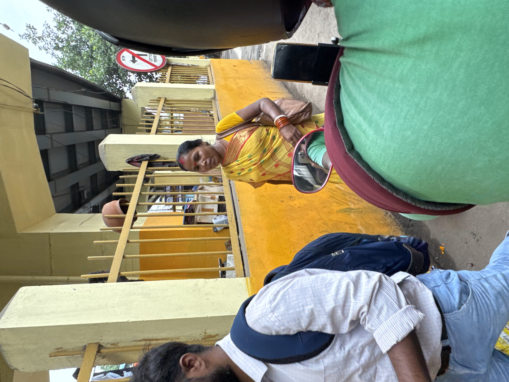In Kolkata, India, on an overcast day, a woman in a yellow sari stands in front of a vibrant orange wall, partially obscured by a yellow metal fence. She wears bangles on her wrist and a bindi on her forehead. To her left, a man wearing a light-colored, checkered shirt and blue jeans, with a blue backpack, walks away from the camera. In the foreground, a portion of a person on a scooter is visible, with a green shirt and a dark red collar. Behind the woman, a traffic sign with a red circle and a diagonal line, indicating "LEFT TURN PROHIBITED," is mounted on a pole. The scene is under an elevated structure, possibly a flyover, with visible metal beams and wiring.