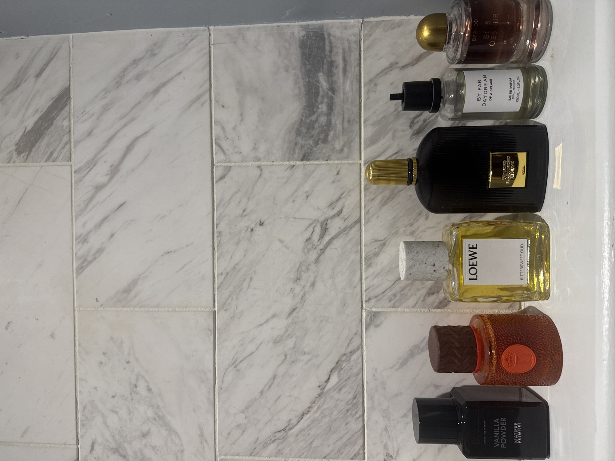 The image shows a collection of six perfume bottles arranged in a vertical row on a surface covered with white marble tiles. The bottles are of various shapes, sizes, and colors, and their labels indicate different brands and scents. From top to bottom, they include: a Byredo Boss Obsur perfume, a By Far Daydream of a Splash perfume, a Tom Ford Black Orchid perfume, a Loewe Bittersweet Oud perfume, a perfume with a round, orange bottle, and a Matiere Premiere Vanilla Powder perfume. The background consists solely of the marble tile surface. The scene does not depict a location in London or feature any people or actions.
