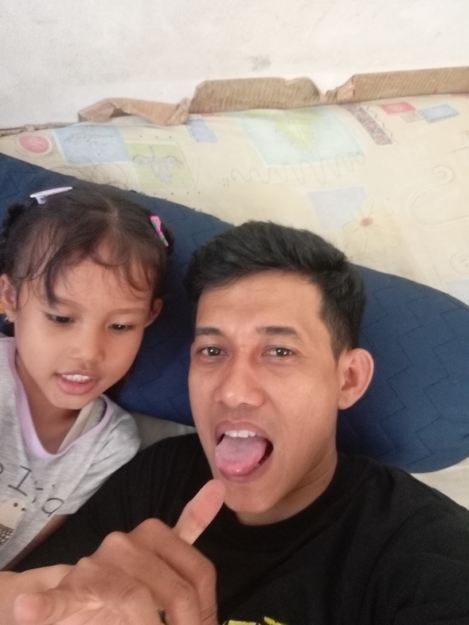 A close-up selfie shows a man and a young girl. The man is on the right, looking towards the camera with his tongue sticking out. He is wearing a black t-shirt. The girl is on the left, looking down and to the left, with her mouth slightly open. She is wearing a grey t-shirt with some text and an image on it. Both are positioned in front of a blue pillow and a patterned blanket. The background appears to be a wall with some decorative elements. The overall impression is a casual, indoor setting. The image was taken in Batam, Indonesia.
